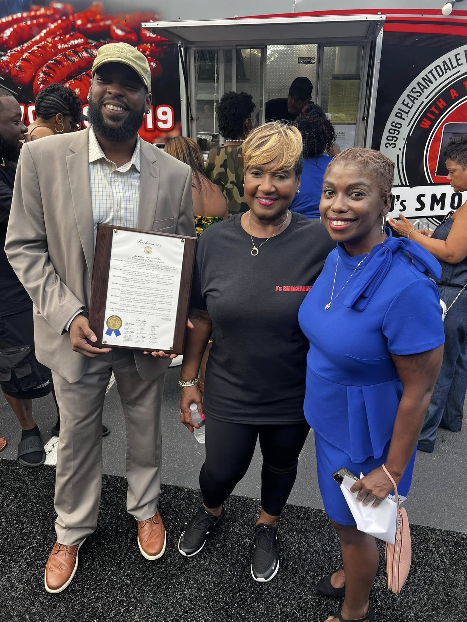 Three people standing outdoors, smiling, with a fire truck and crowd in the background. One person holds a framed certificate.