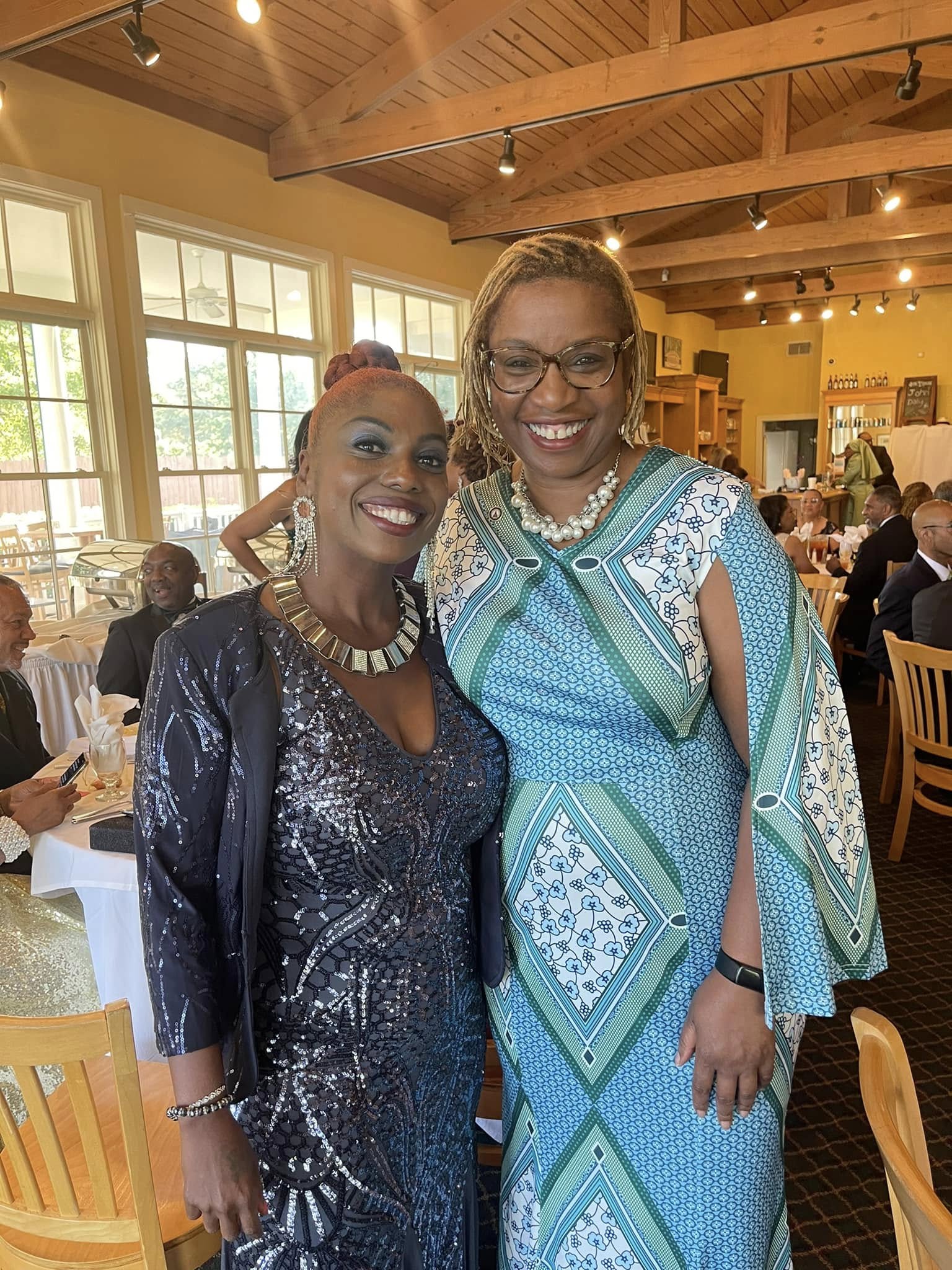 Two women smiling indoors at a social event, one wearing a dark shiny dress and the other in a colorful patterned dress, with a wooden ceiling and large windows in the background.