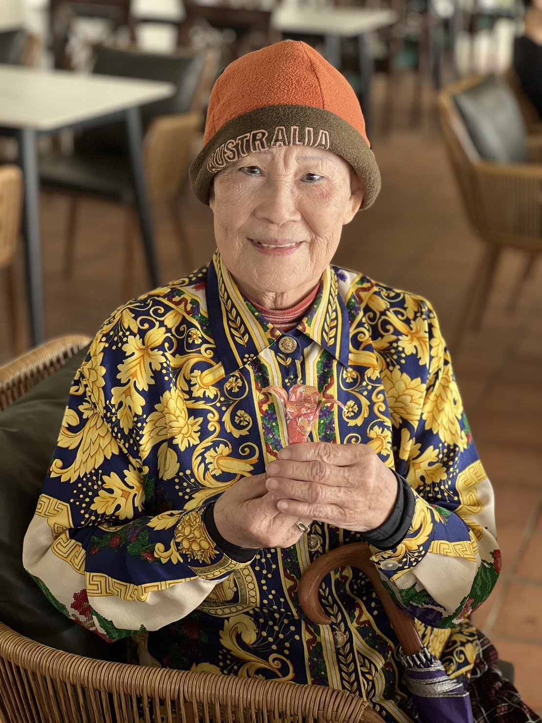 Older woman wearing a colorful patterned shirt, orange and gray beanie, smiling with hands clasped, in a room with chairs and tables.