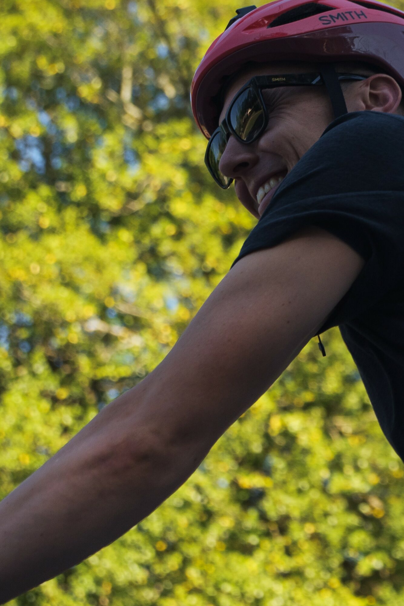 Person wearing a pink helmet and sunglasses smiling outdoors with green trees in background.