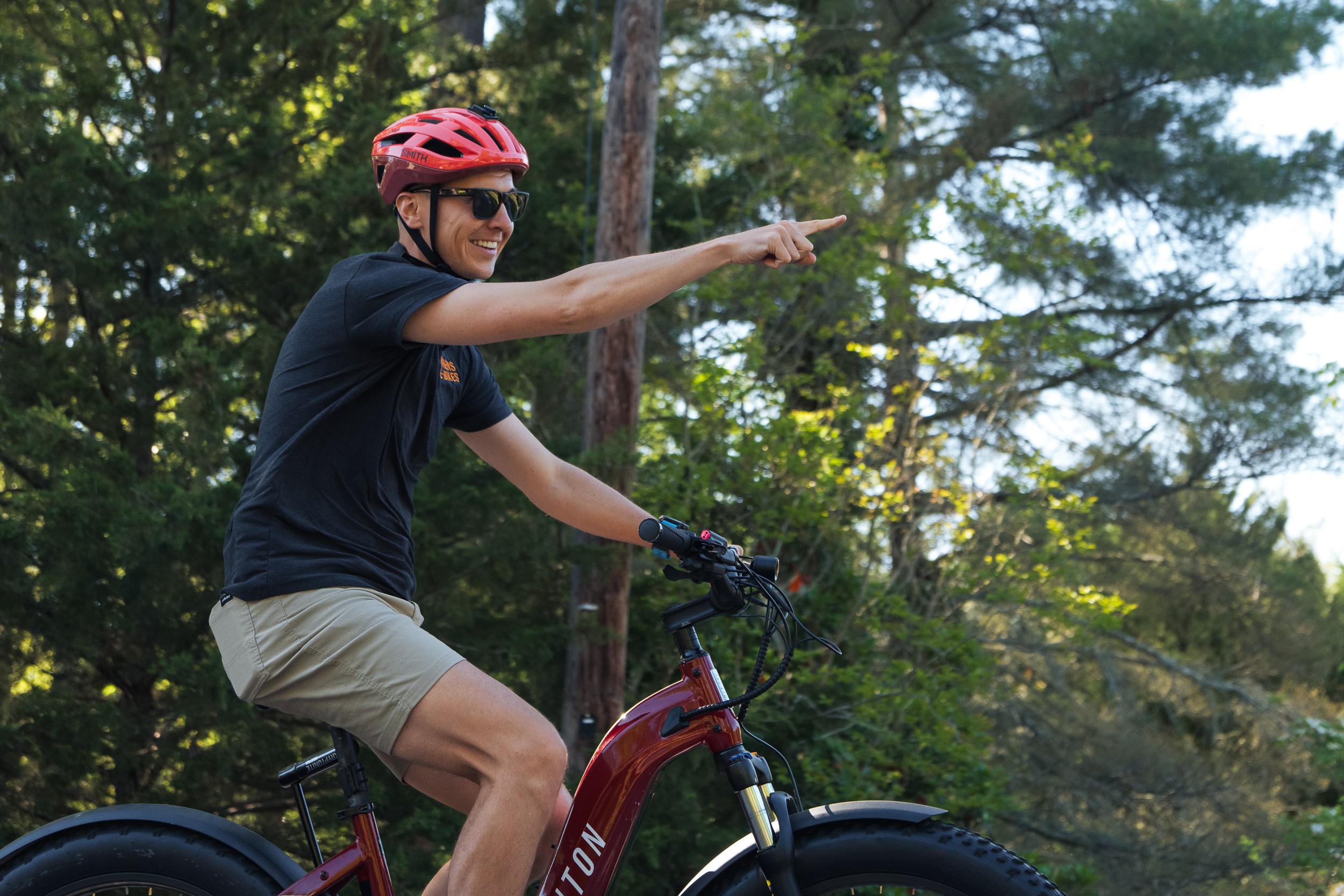 Person wearing a red helmet and sunglasses, riding a bicycle outdoors, pointing to the right, surrounded by trees.