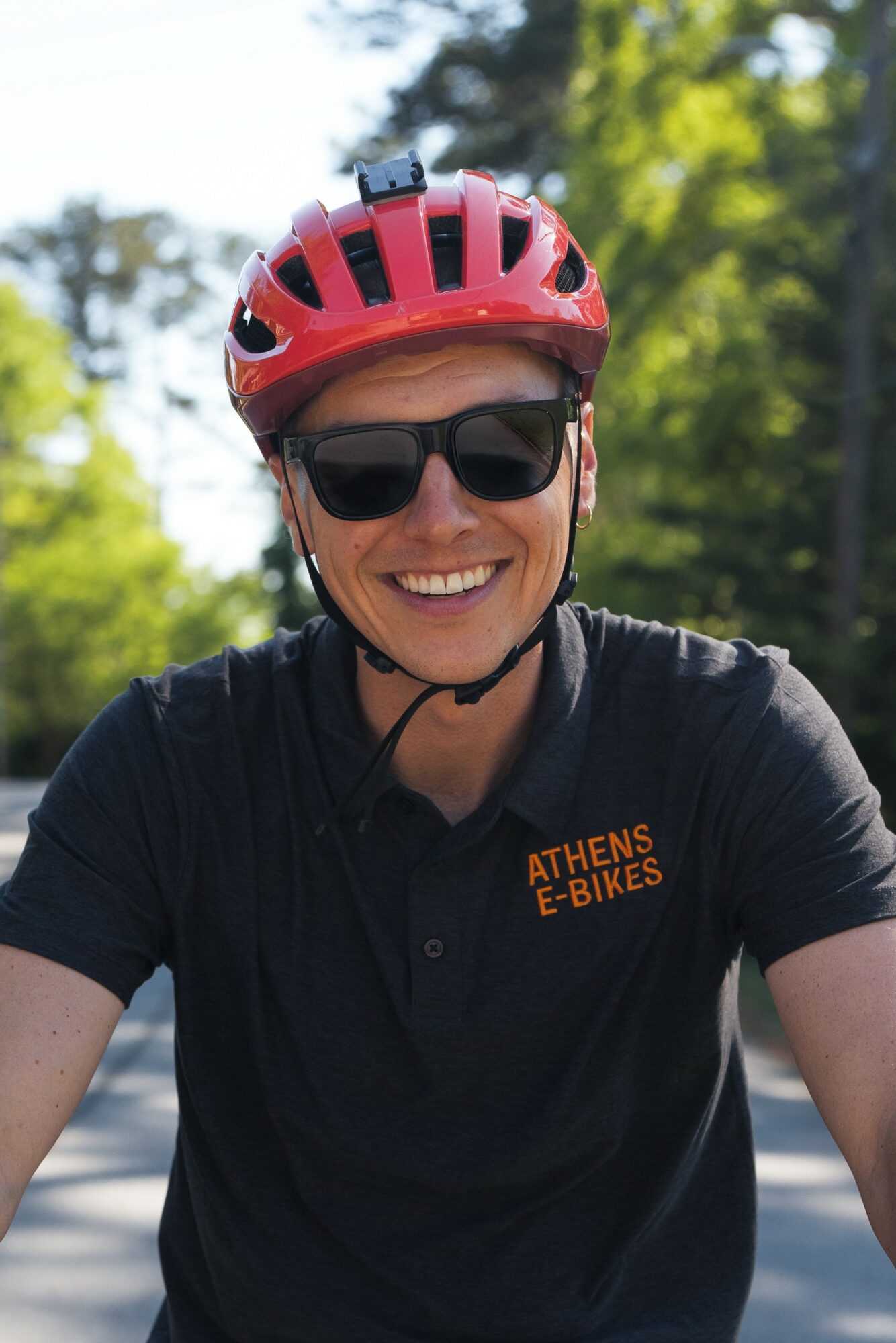 Smiling person wearing a red helmet and sunglasses outdoors, with trees in the background.