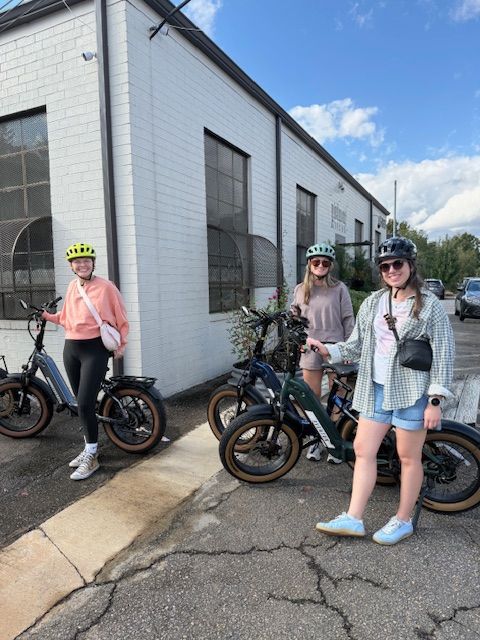 Two children with helmets and a woman with a bicycle on a cracked sidewalk outside a white building under a blue sky.