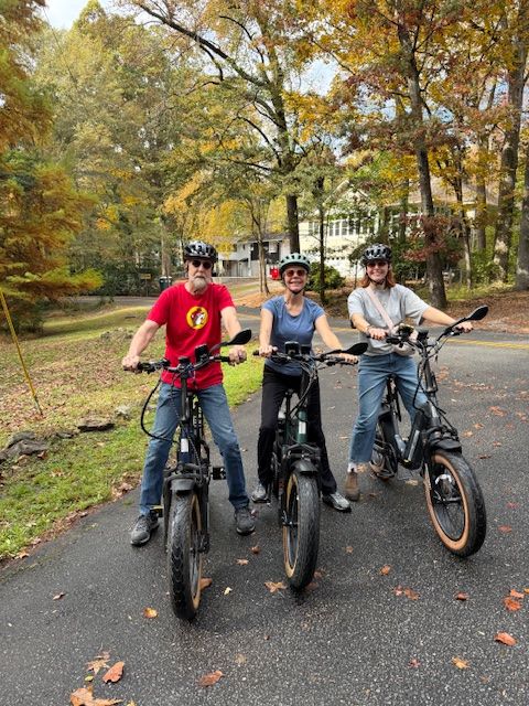Three people with bicycles on a paved path in a park with trees and fallen leaves.