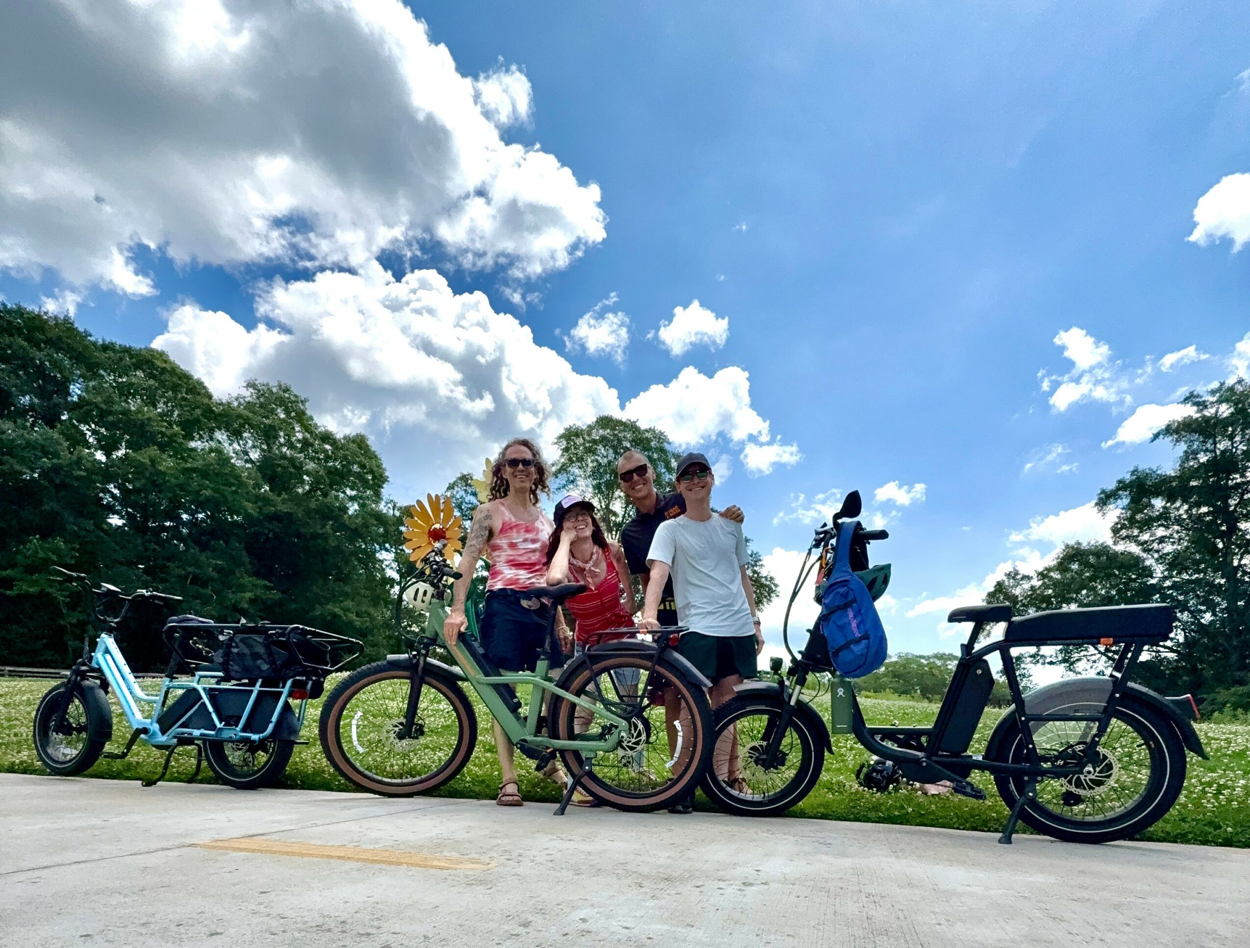 Group of four people with bicycles outdoors on a sunny day, trees and clouds in the background.