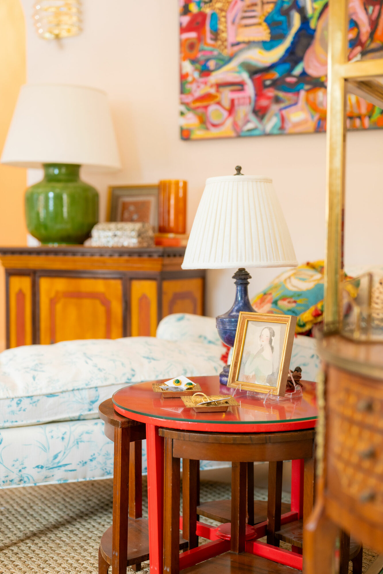 Living room with a wooden side table, lamps, colorful artwork, and a bed with floral bedding.