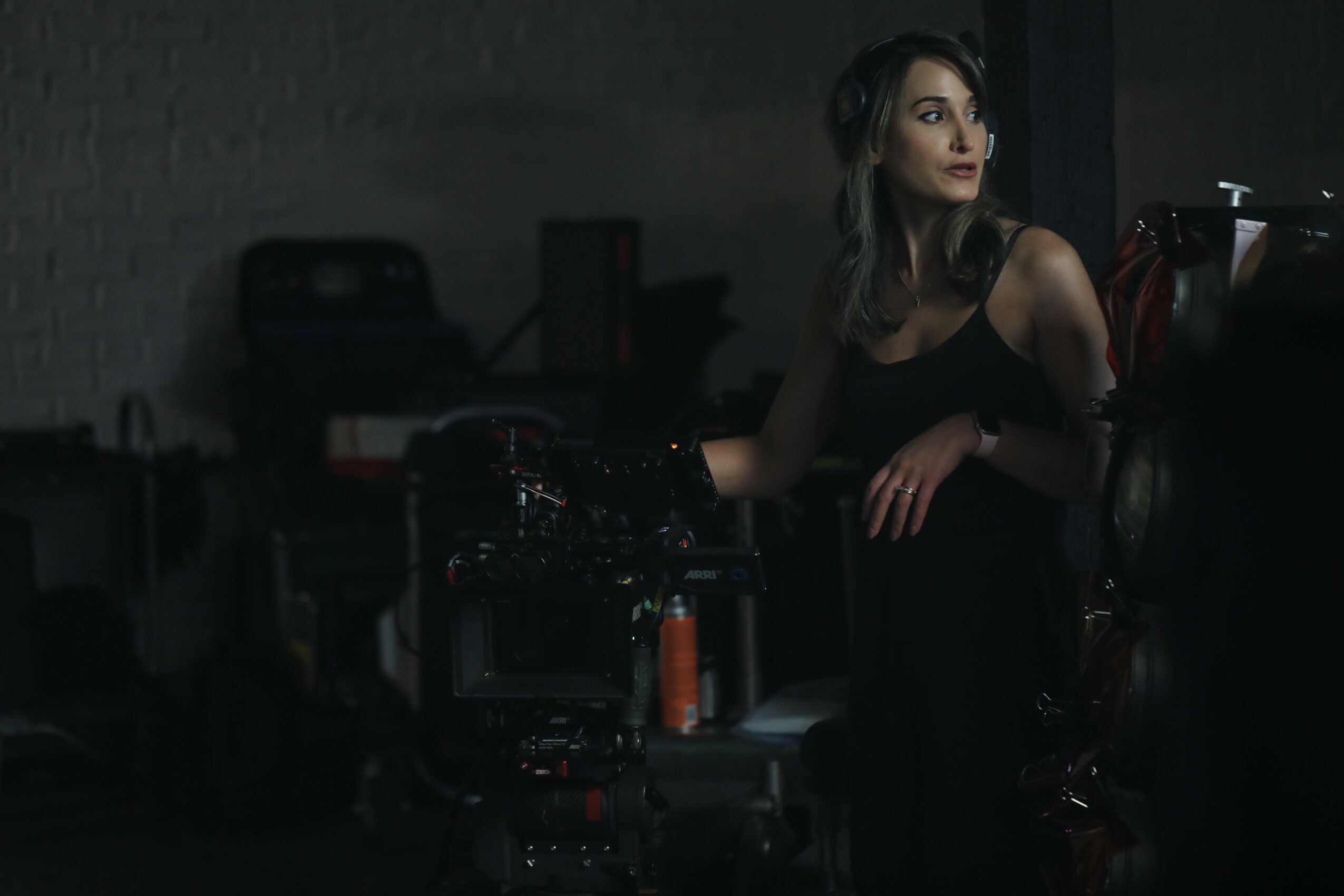Woman with long hair in a dark room, sitting and looking to the side, with equipment and a dark background.