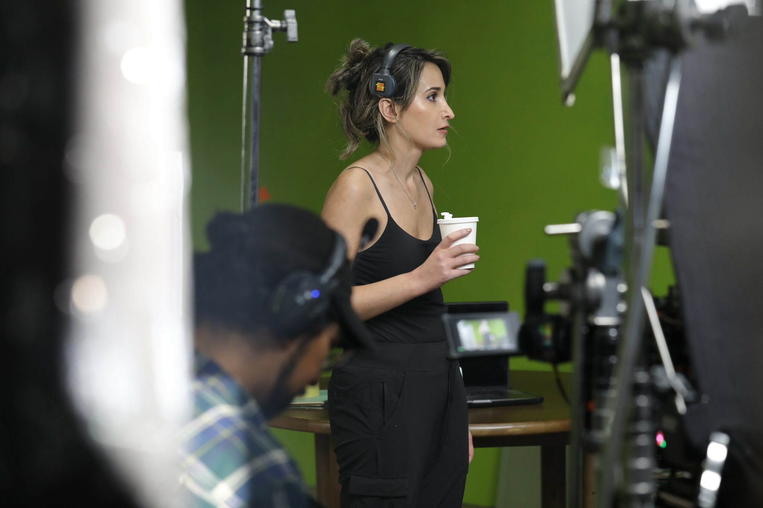 Woman with headphones holding a cup, surrounded by filming equipment, in front of a green screen.