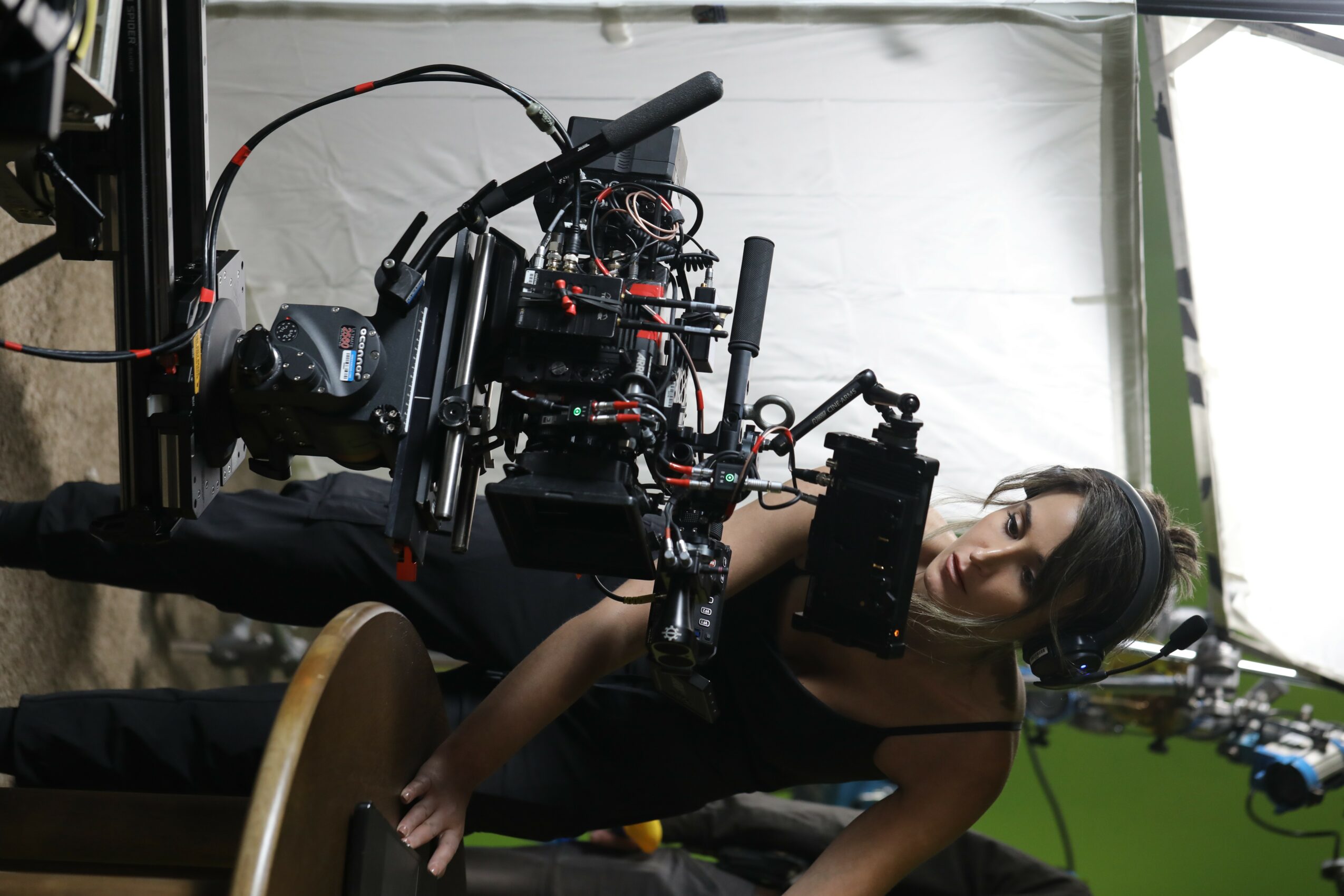 Woman operating a large camera or filming equipment indoors, with a white wall and green background behind her.
