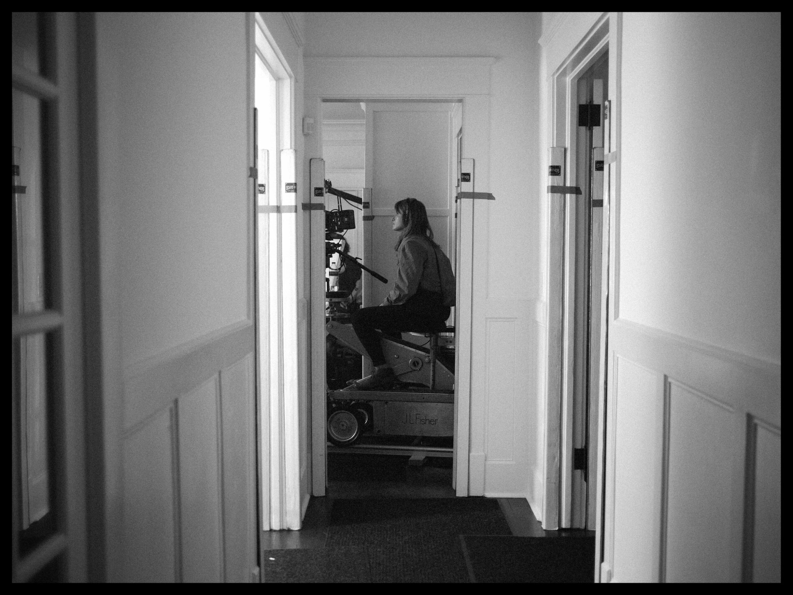 Person sitting at a desk in a room, viewed through a doorway in a hallway, black and white photo.