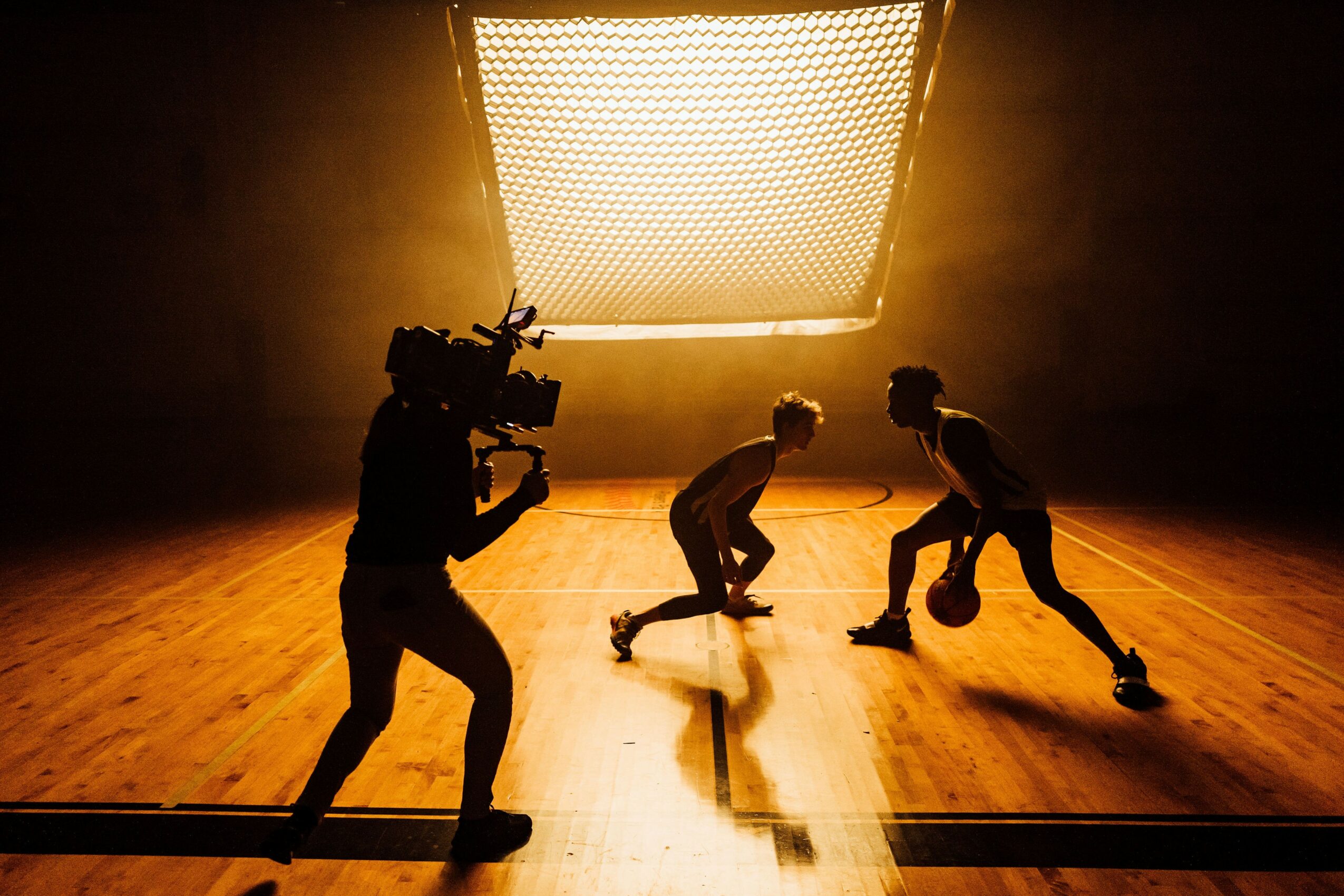 Two children playing basketball on a wooden court with a bright light overhead, and a person filming them.