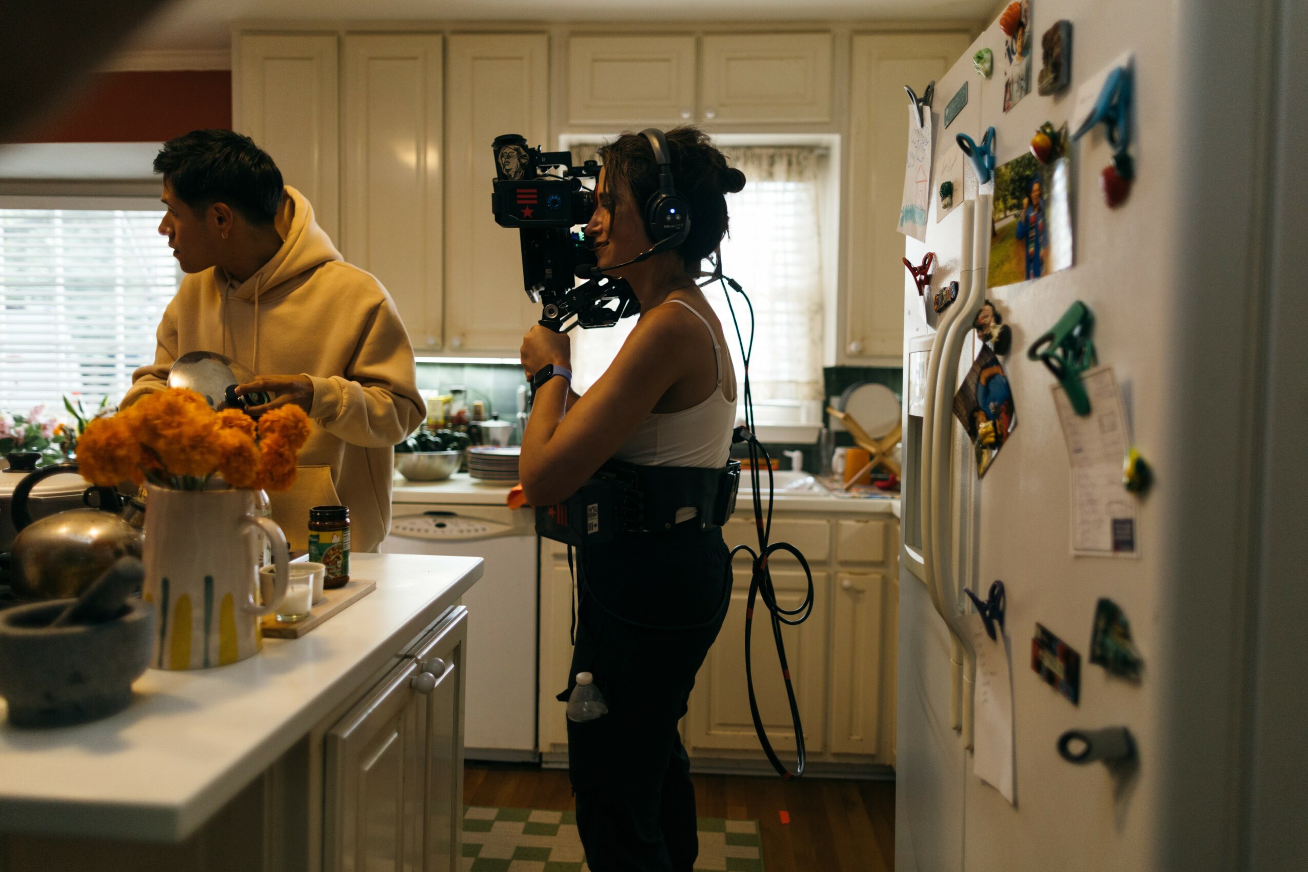 Two people in a kitchen, one filming with a camera, the other preparing food, with colorful magnets on the fridge.