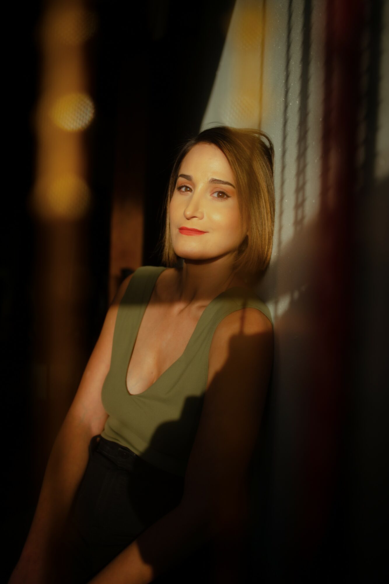 Woman with shoulder-length hair and a sleeveless top, standing near a window with curtains, illuminated by warm light.