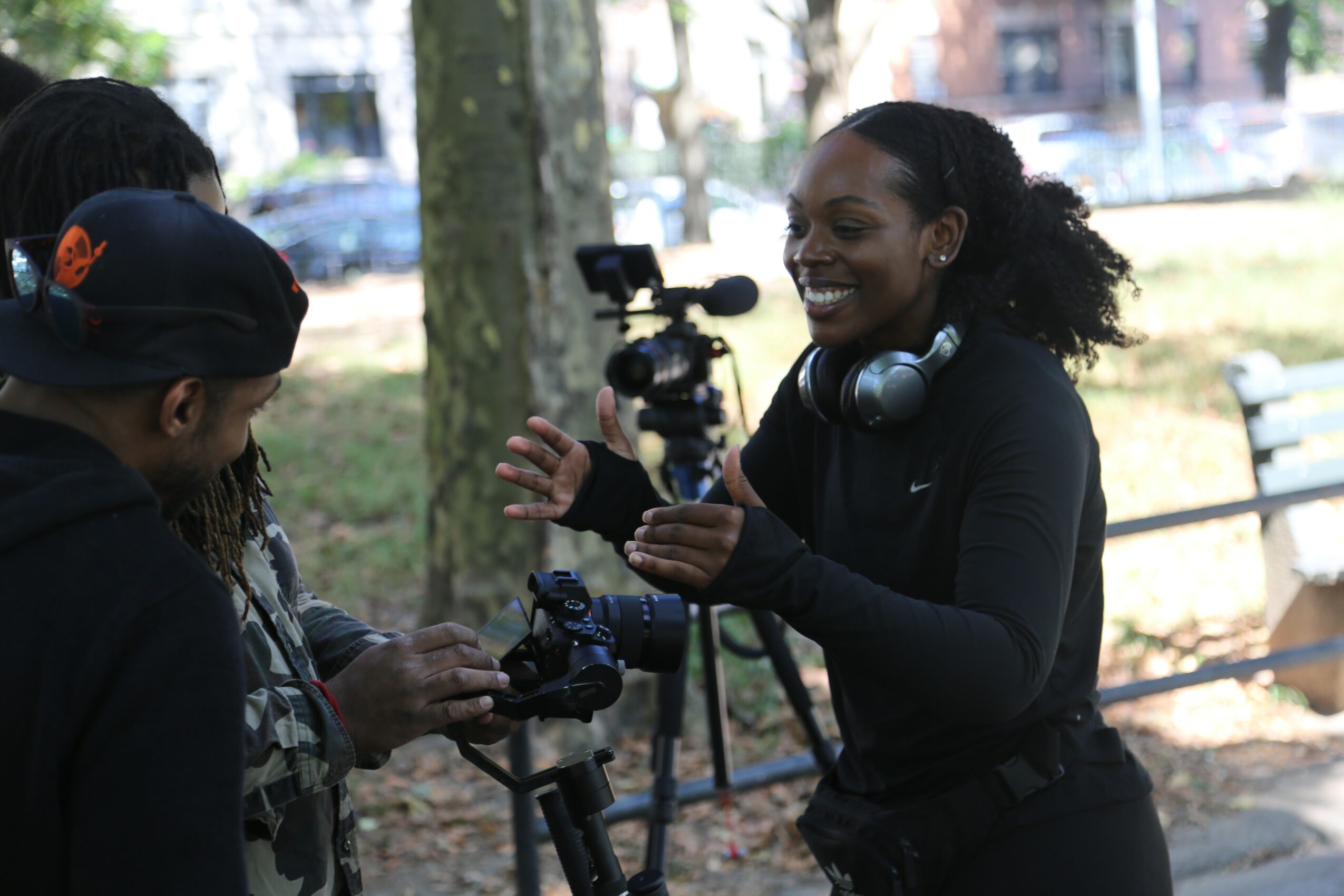 Three women outdoors, one with headphones, smiling and talking, with a camera on a tripod, trees and benches in background.