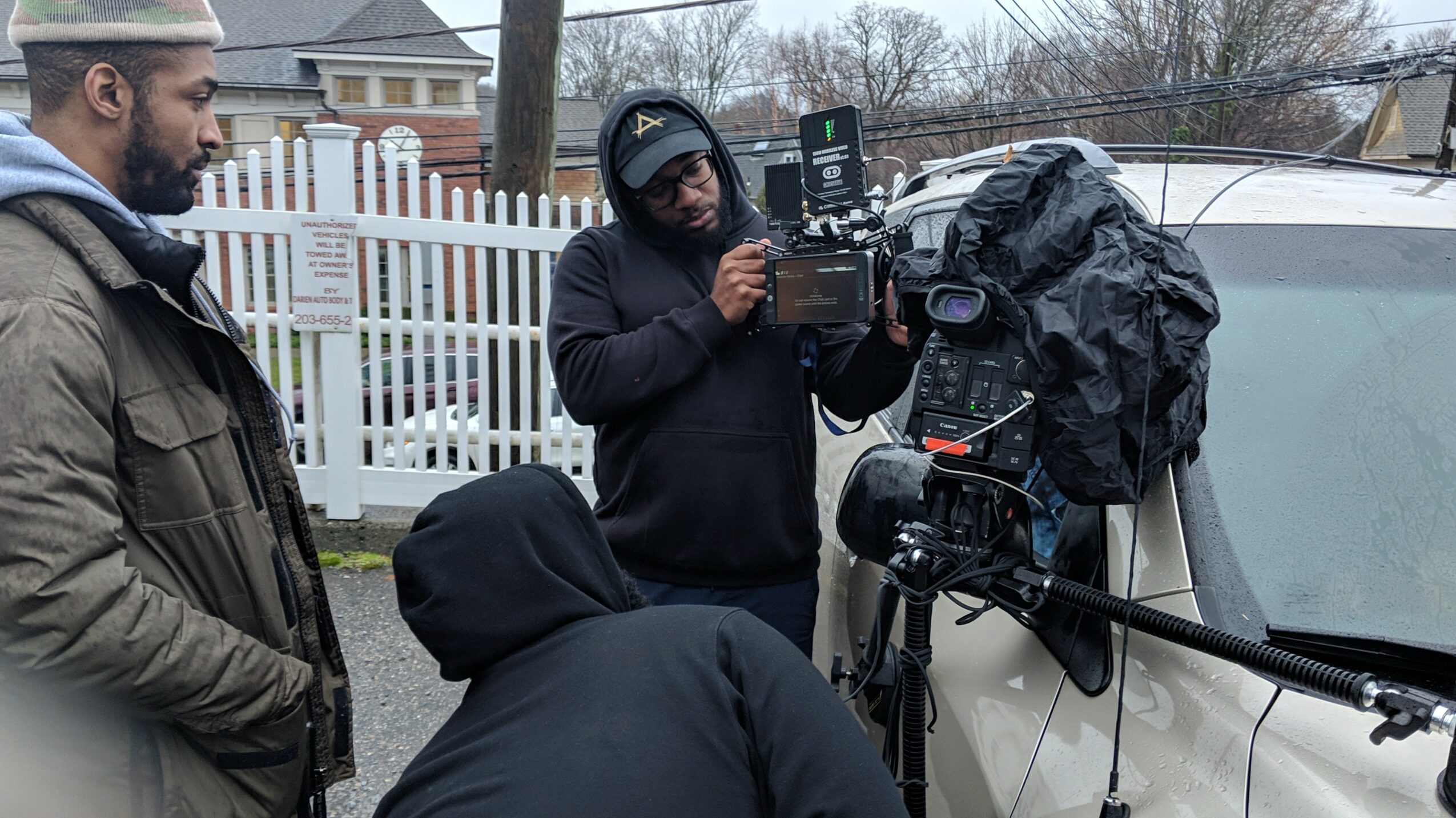 Three people outdoors near a white vehicle, with one person operating a camera and another observing, in a residential area.