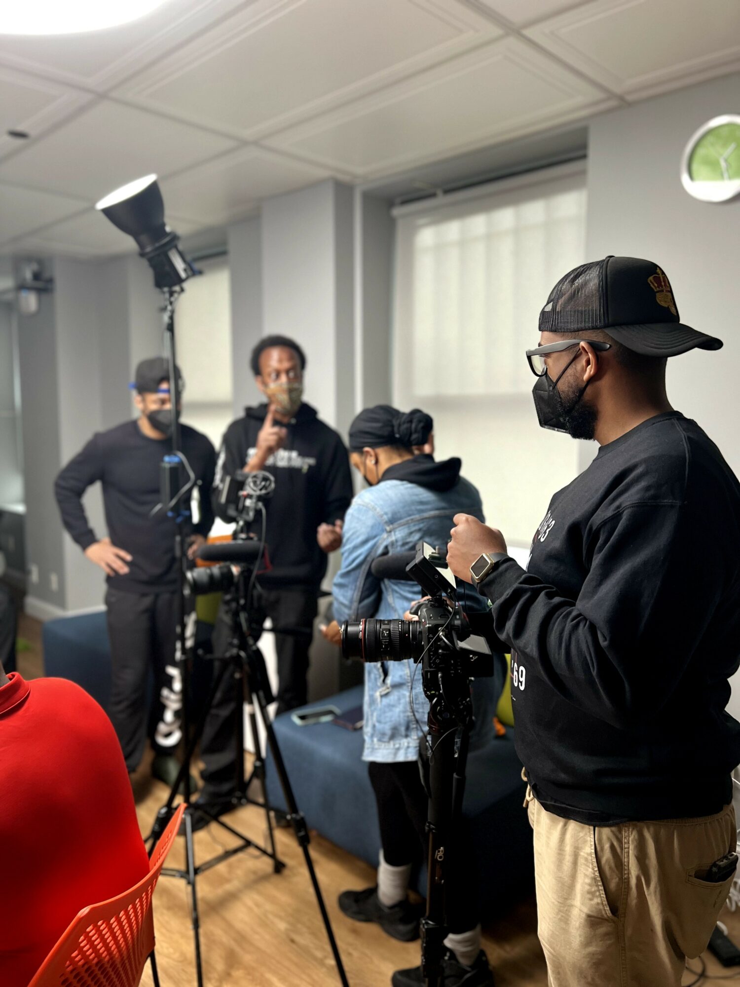 Group of people in a room with camera equipment, some wearing masks, engaged in a discussion or filming activity.