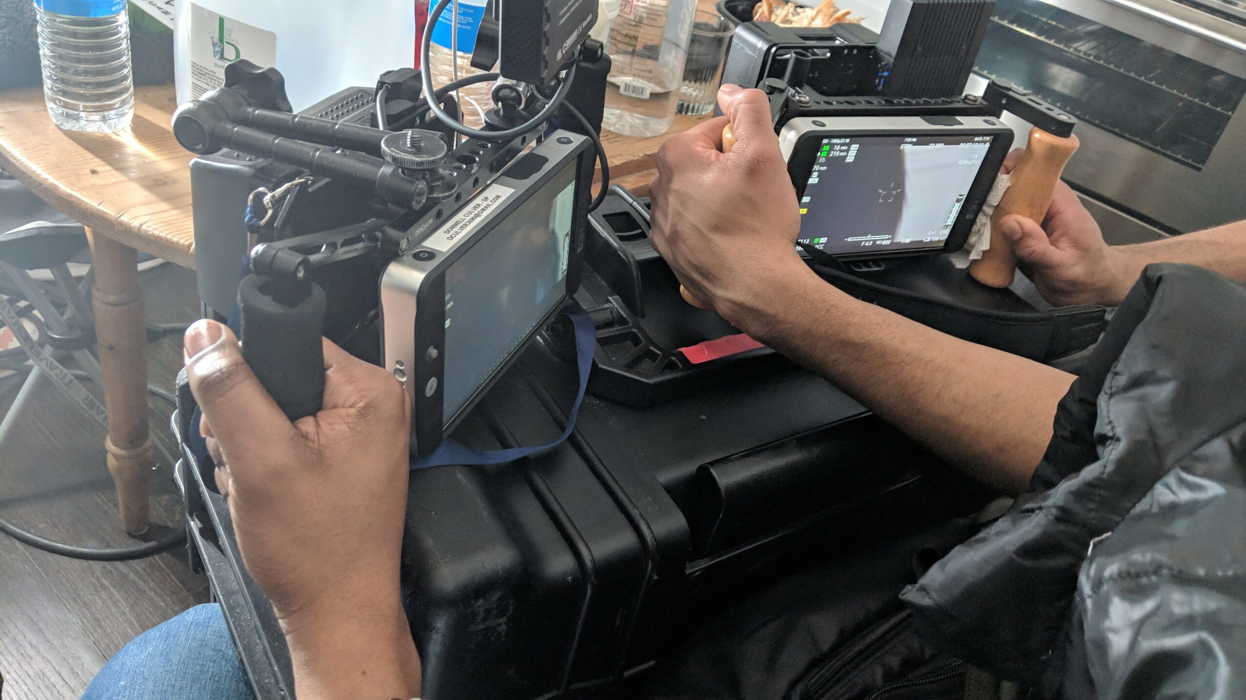 Two people hold cameras and tablets, working on a black case with electronic equipment on a wooden table.