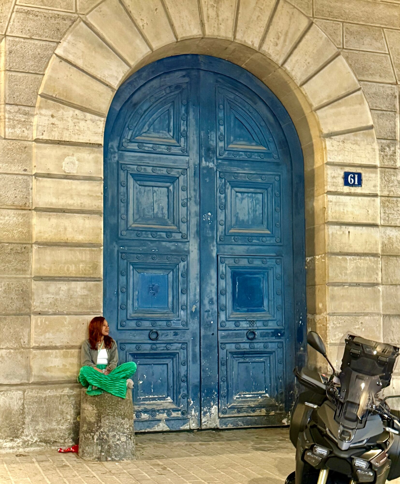 Person with red hair sitting on a stone block in front of a large blue arched door, with a motorcycle nearby.