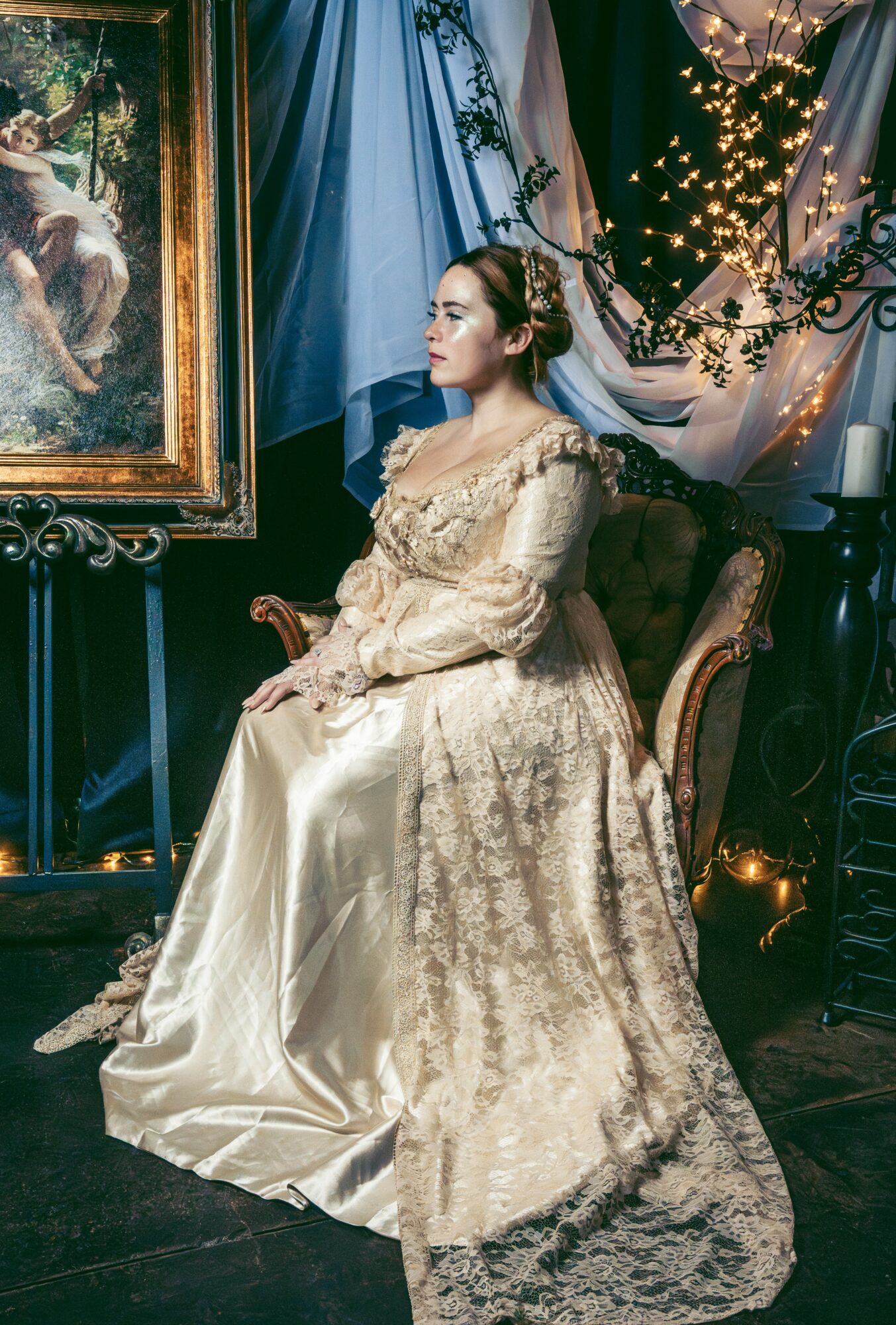 Woman in a cream-colored gown sitting on a vintage chair in a decorated room with a painting and string lights.