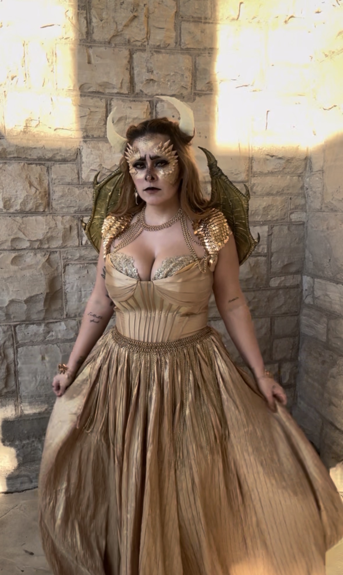Woman in gold dress with makeup and headpiece, standing against a stone wall, illuminated by sunlight.