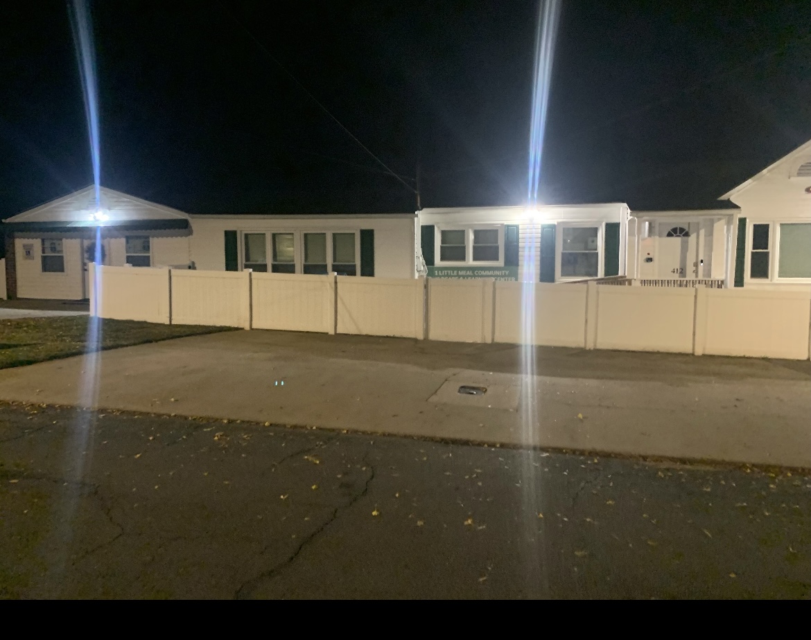 Night scene of a row of houses behind a white fence, illuminated by streetlights, with a sidewalk in front.
