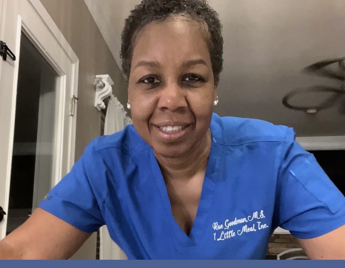 Woman with short curly hair wearing blue scrubs, smiling, in a room with a ceiling fan and window.