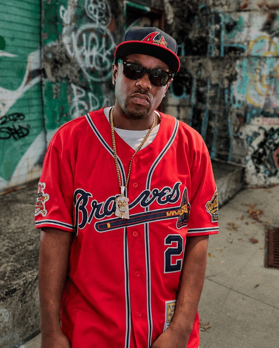 Man wearing sunglasses, a red baseball jersey, and a black cap with a red logo, standing outdoors with graffiti background.