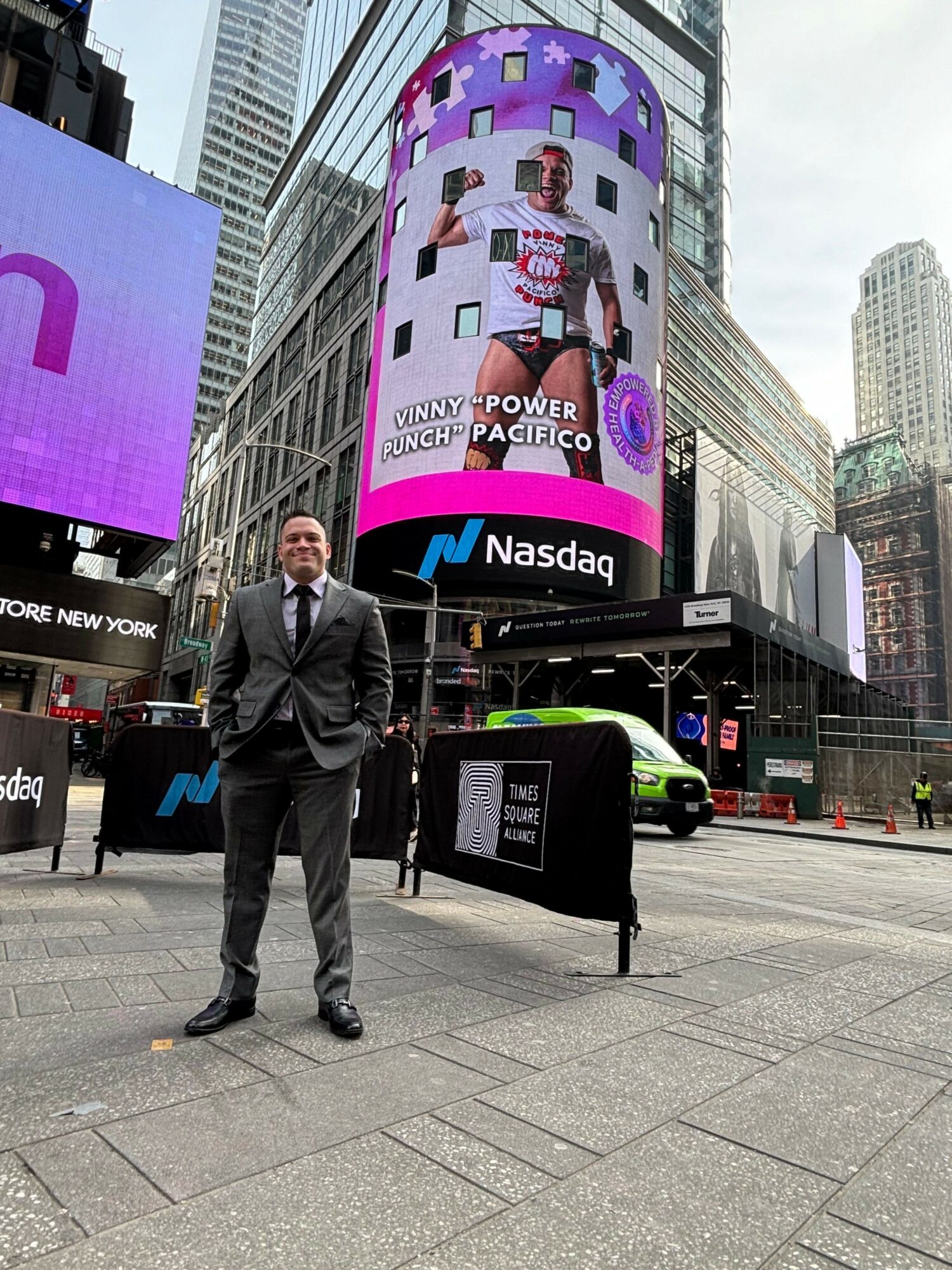 Man in suit standing on sidewalk in front of Nasdaq building with digital billboard and city skyscrapers behind.