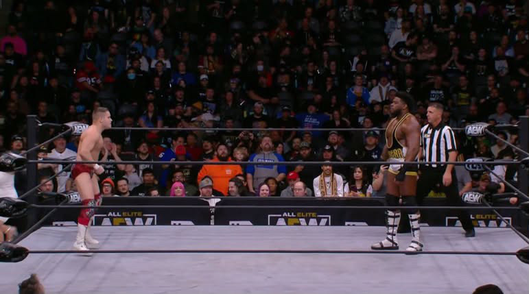 Two wrestlers face each other in a wrestling ring with a referee nearby, and a large audience in the background.
