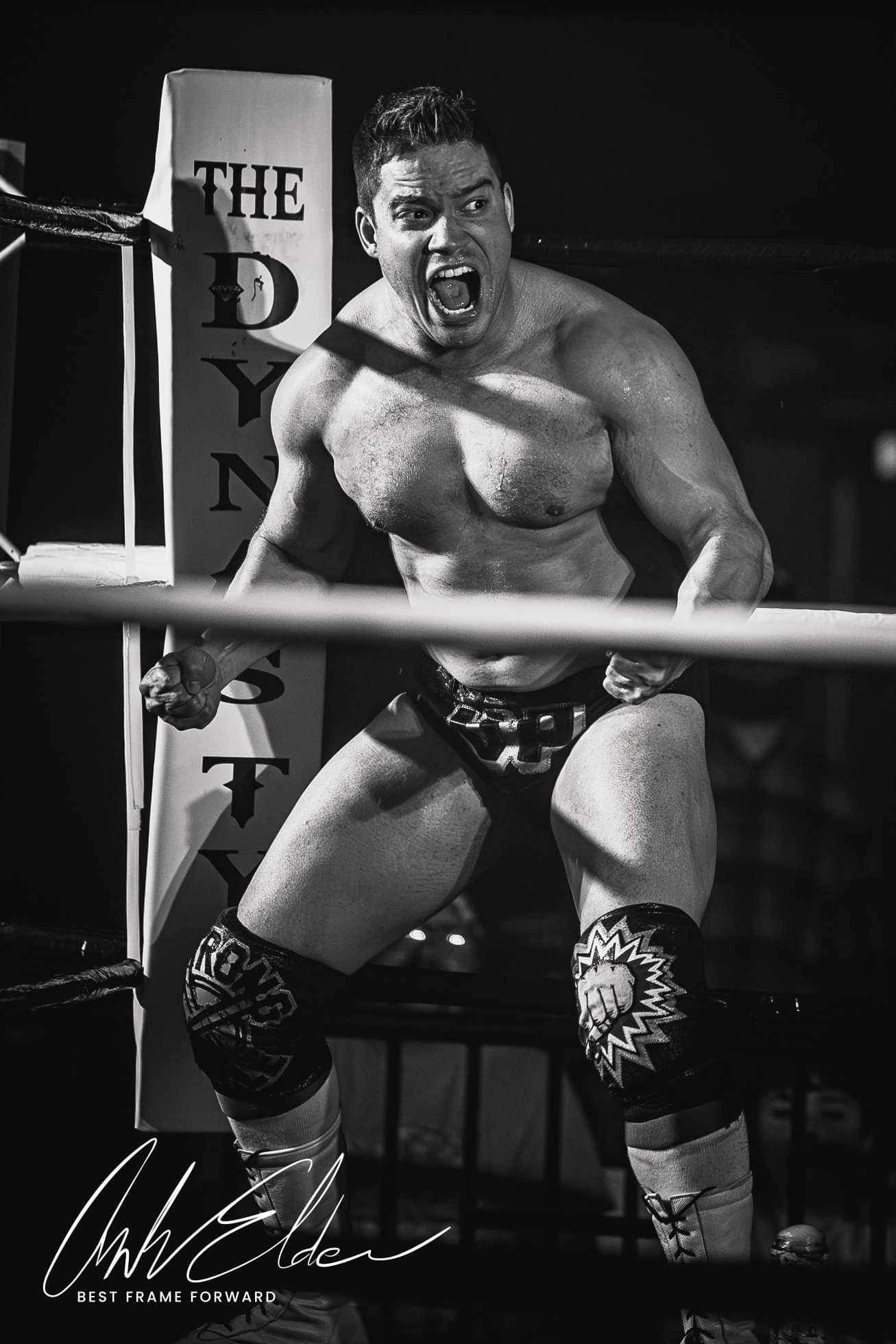 Male wrestler in a ring, shouting with arms flexed, wearing wrestling gear and knee pads, black and white photo.