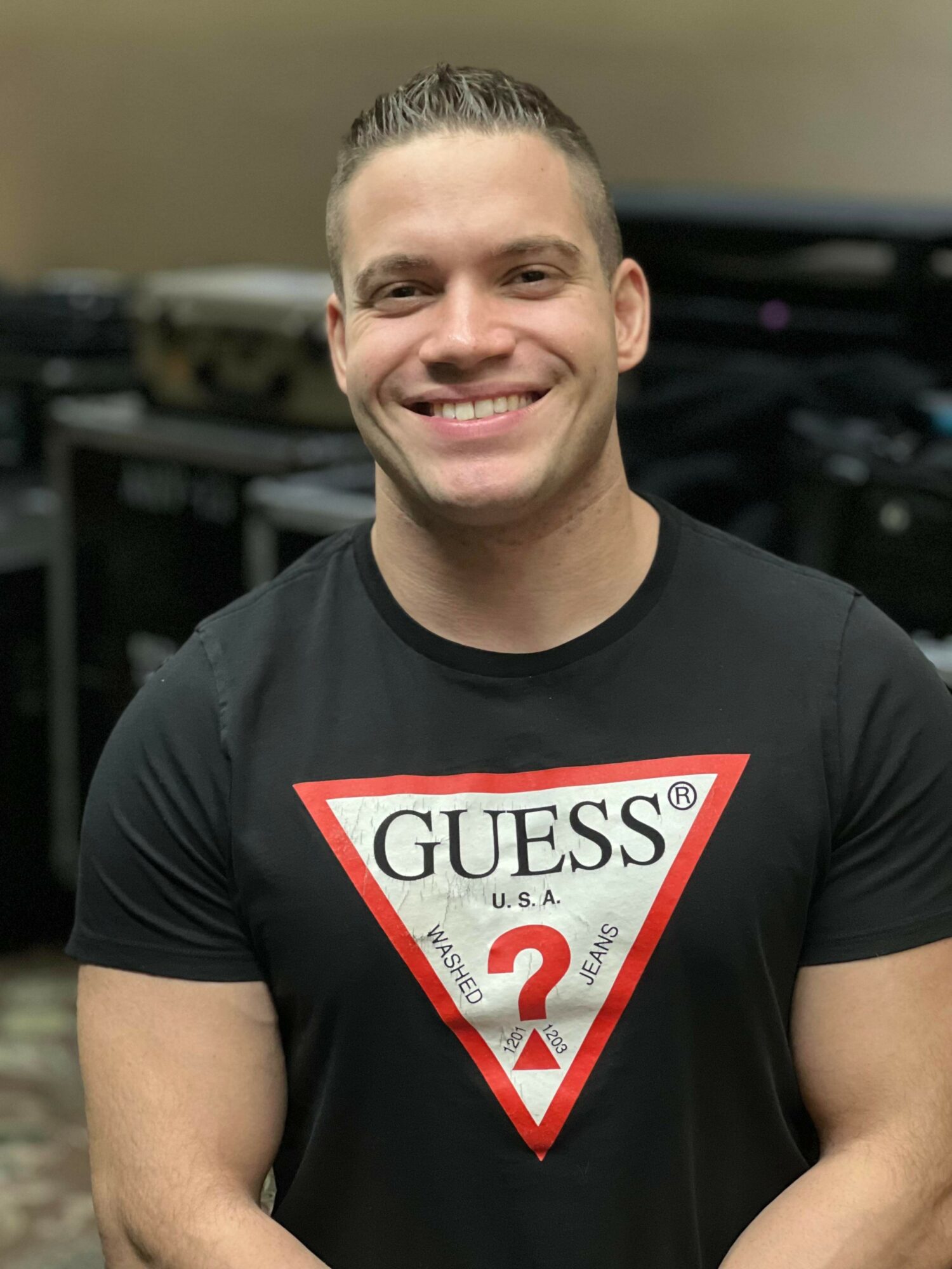 Smiling man with short hair wearing a black Guess T-shirt with a red and white logo, standing indoors.