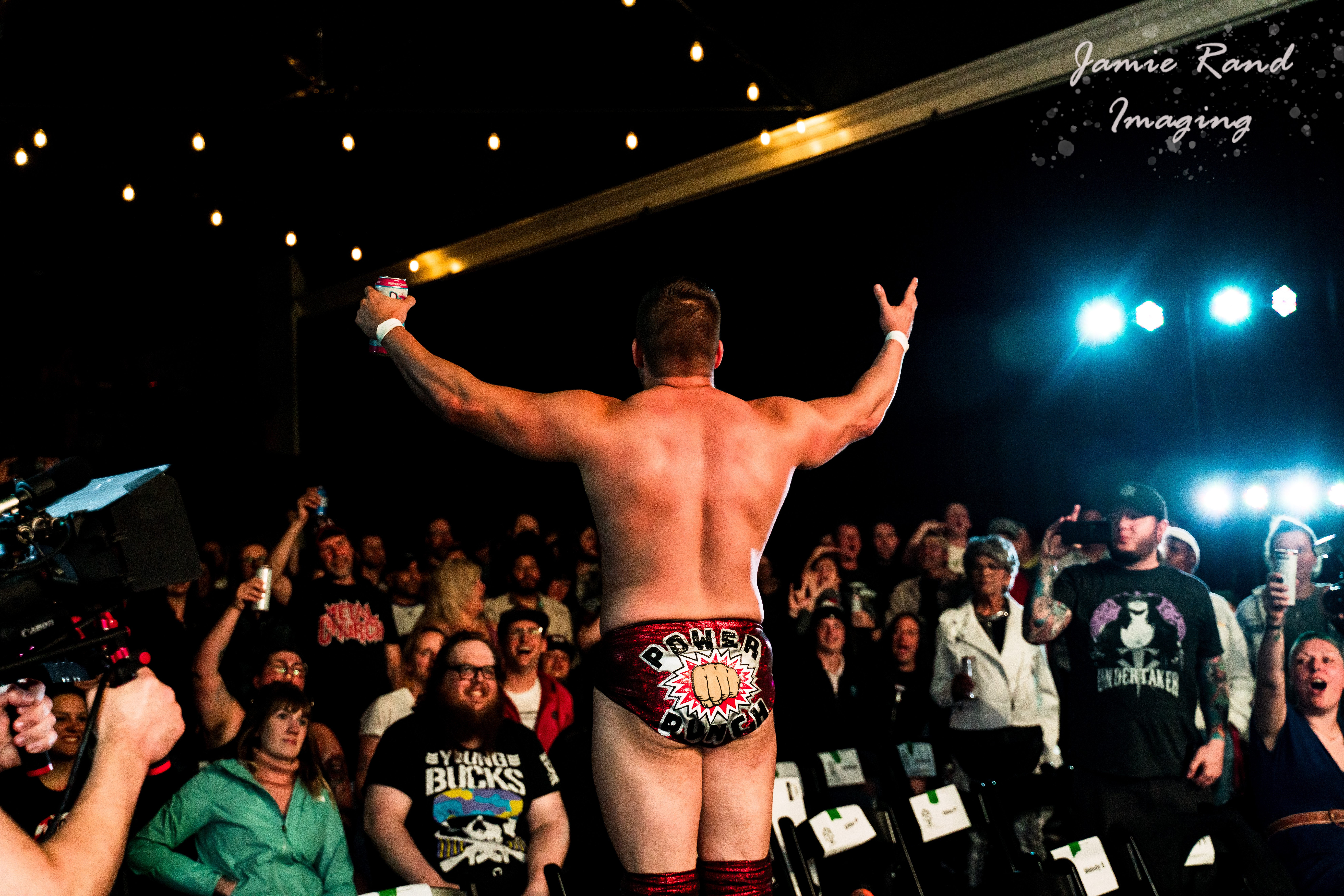 A shirtless wrestler with arms raised in front of a crowd at a wrestling event, bright stage lights overhead.