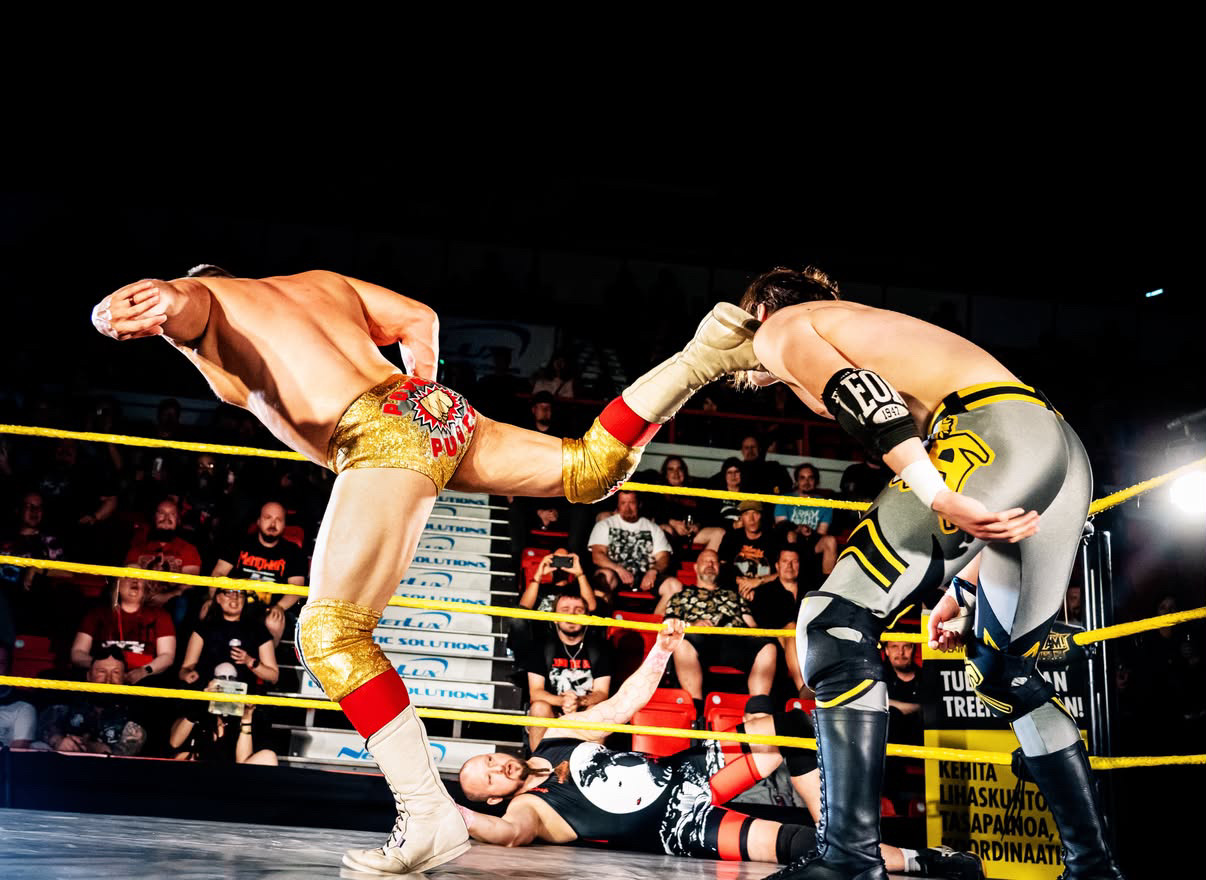 Two female wrestlers in a ring, one delivering a kick to the other's face, with a crowd watching.