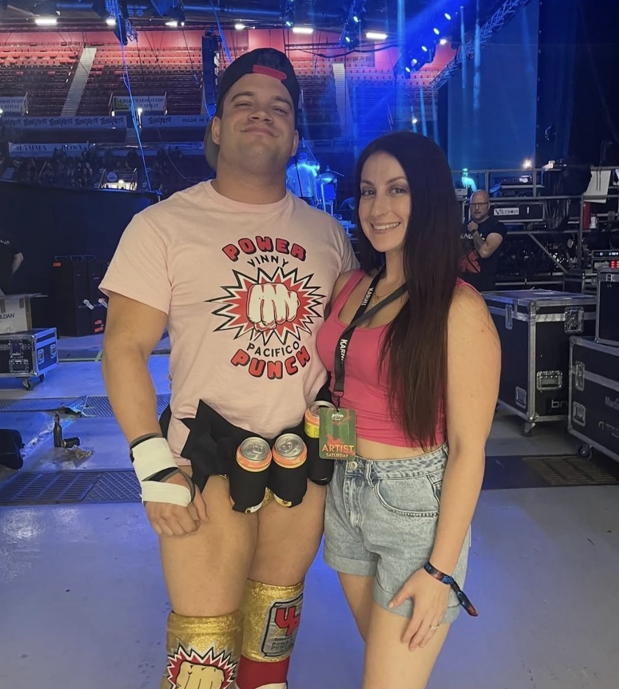 Two people posing on stage, one wearing a pink shirt and shorts, the other in a pink top and denim shorts, stage equipment visible.