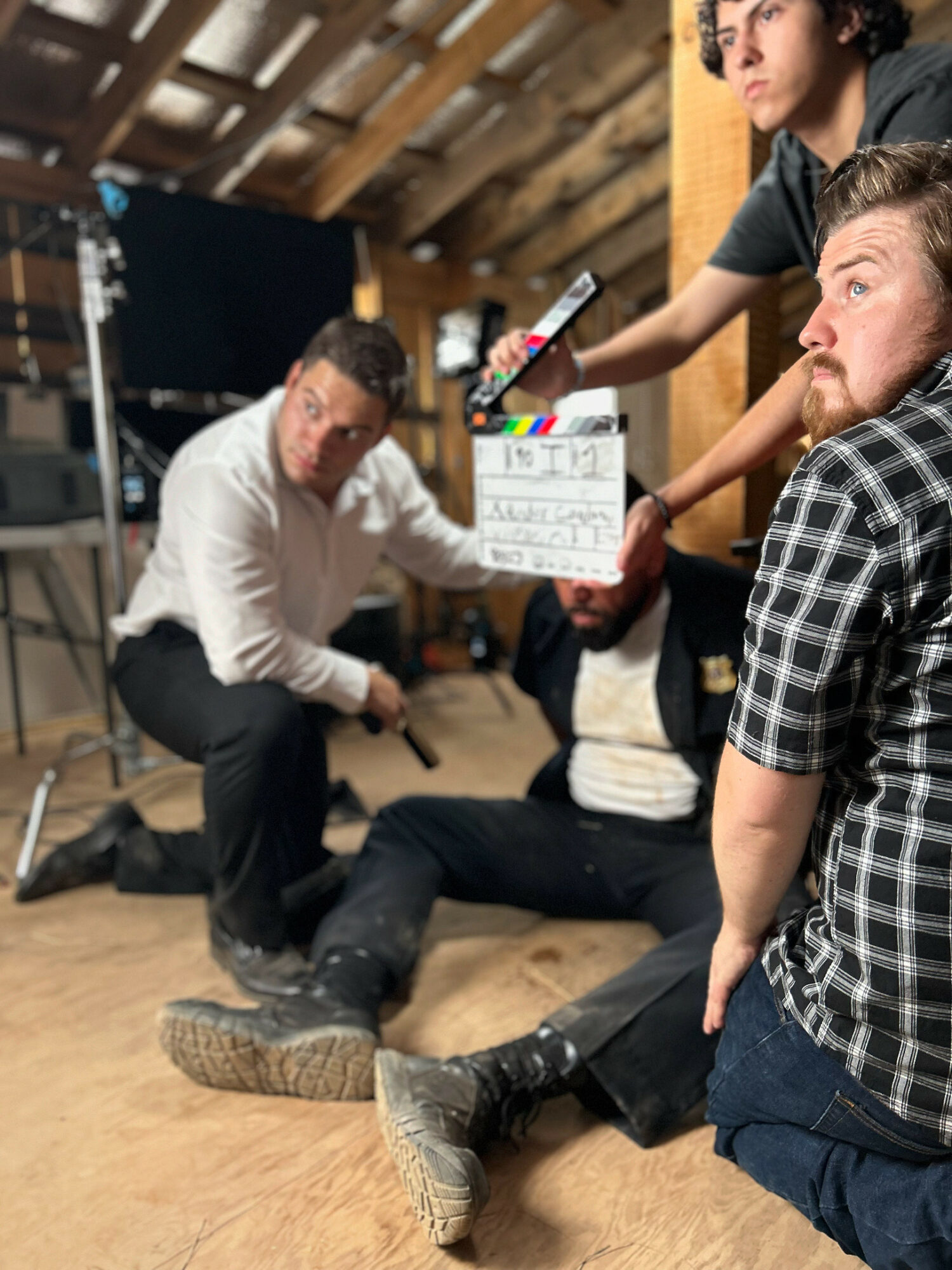 Group of people on a film set, one person sitting on the floor, others standing, holding a clapperboard, in a room with wooden ceiling beams.
