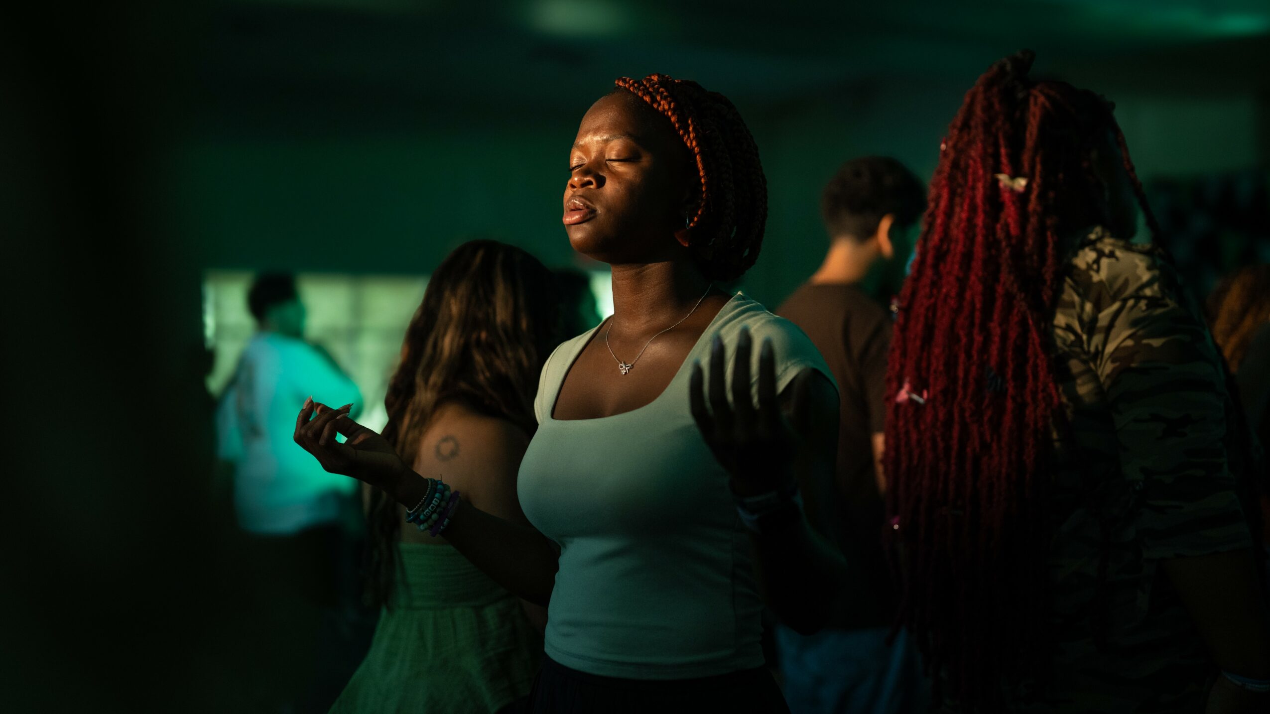 Group of people in a dimly lit room, some with their eyes closed, one woman with braided hair holding a phone.