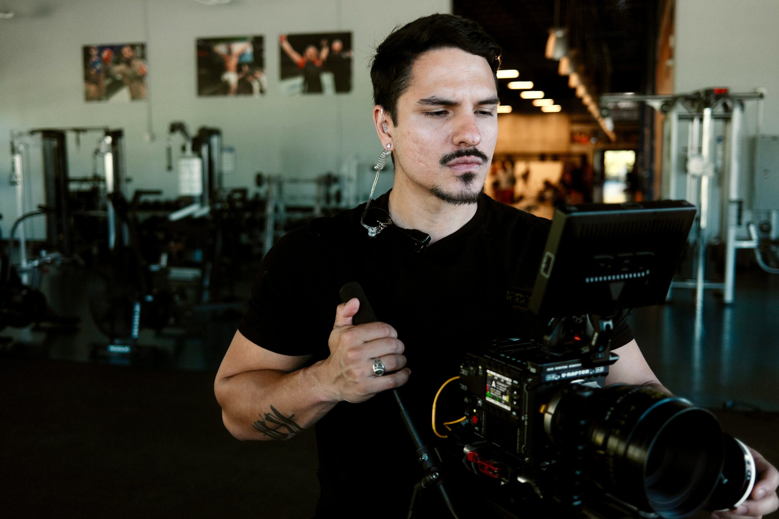 Man operating a professional camera in a gym, with workout equipment in the background.