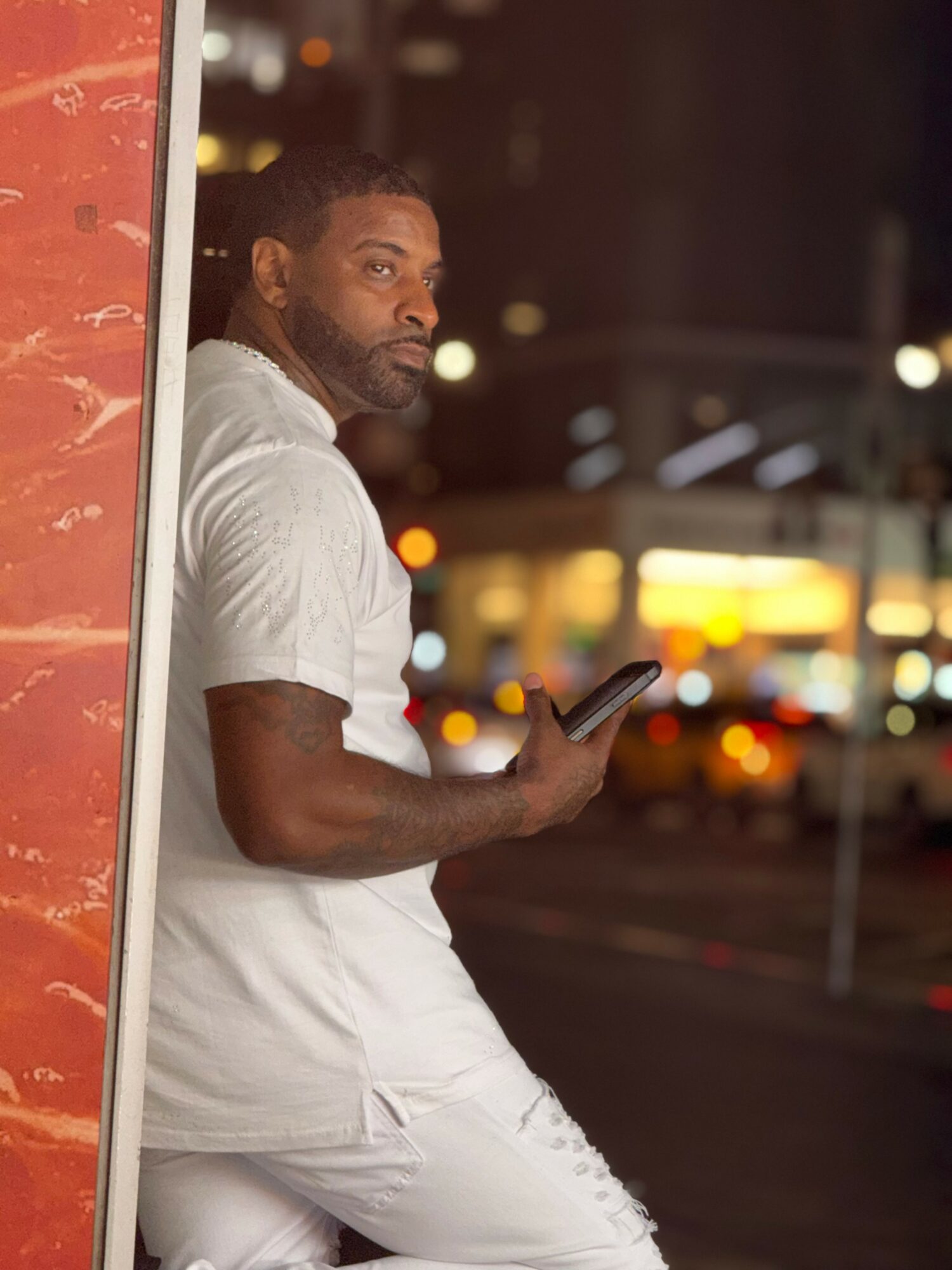 Man in white shirt and pants leaning against a wall, holding a phone, city lights in background.