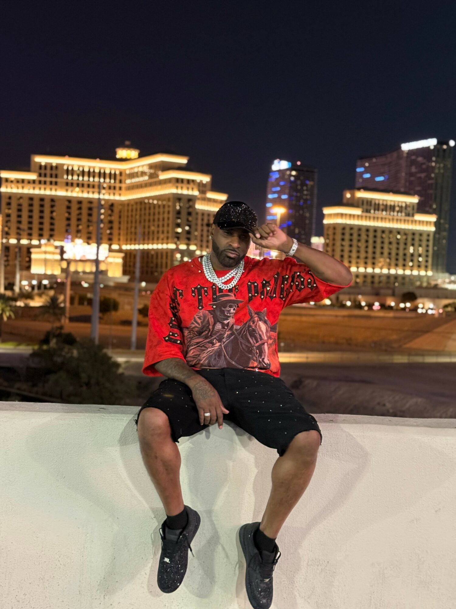 Young man sitting on a ledge at night with city buildings and lights in the background.
