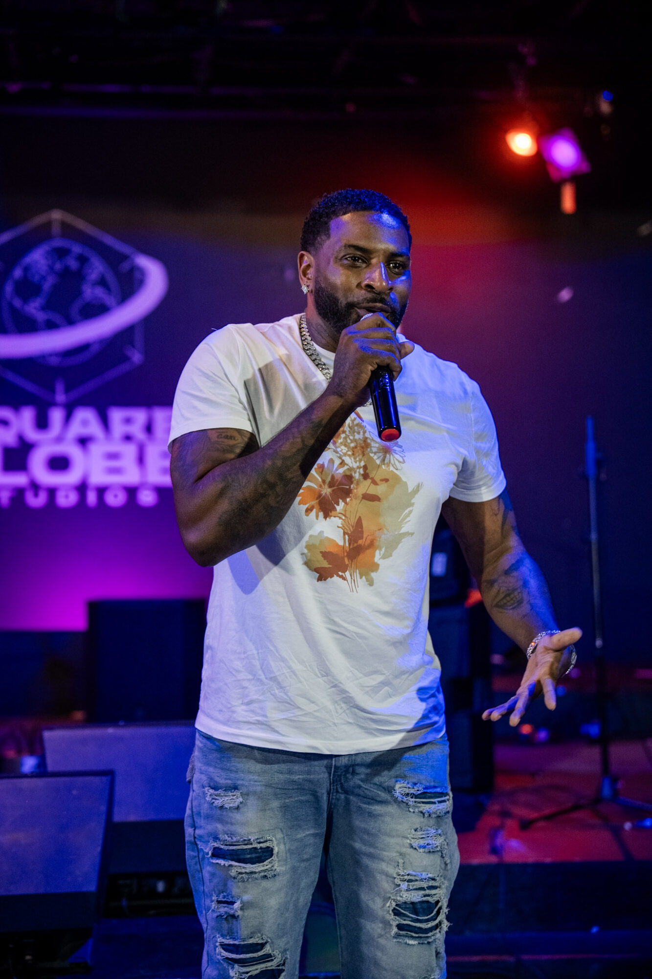 Man holding microphone on stage with purple and red lighting, background shows a logo and text, wearing a white T-shirt and jeans.