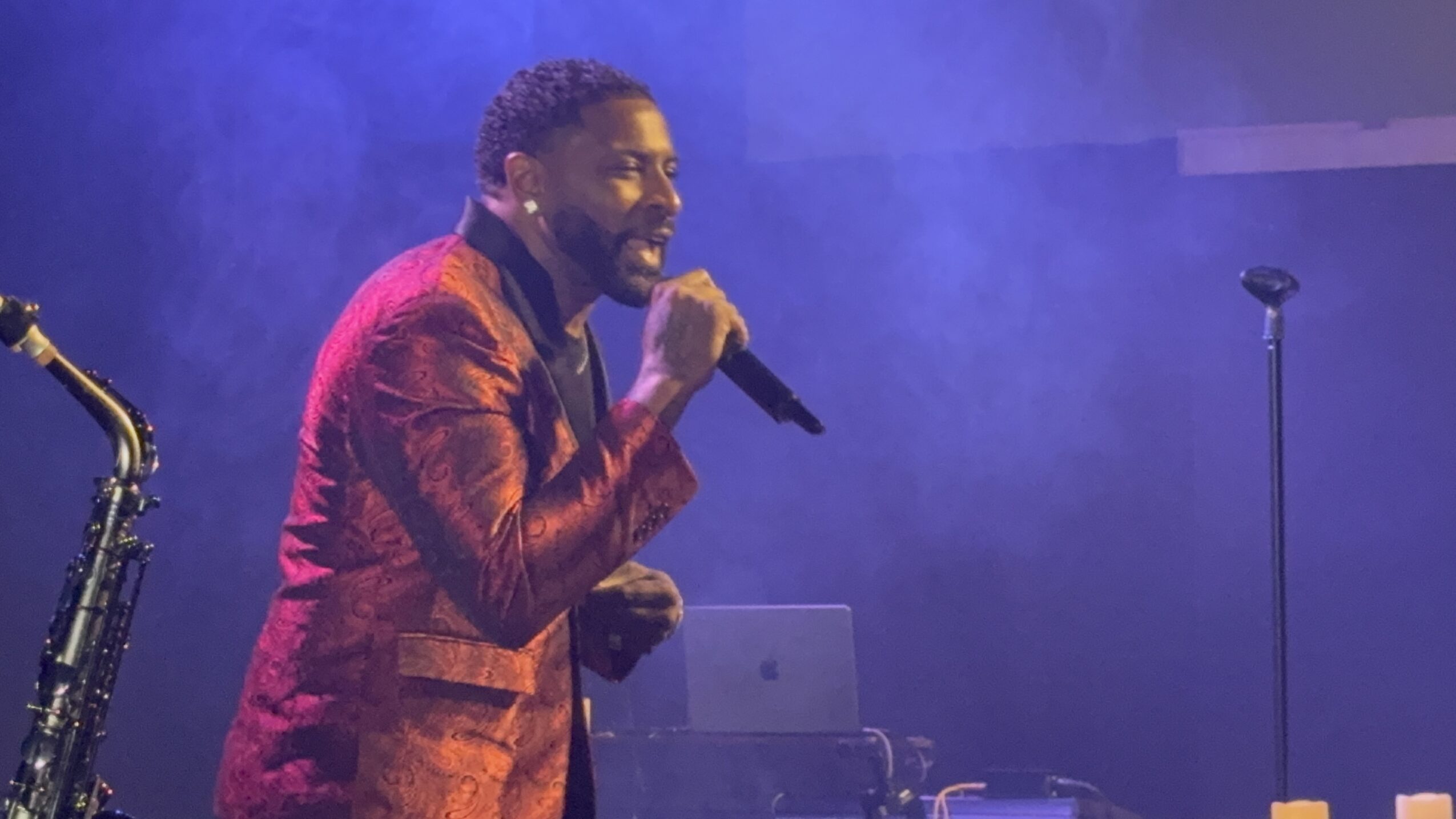 Person singing into a microphone on stage with purple lighting, wearing a red patterned jacket.