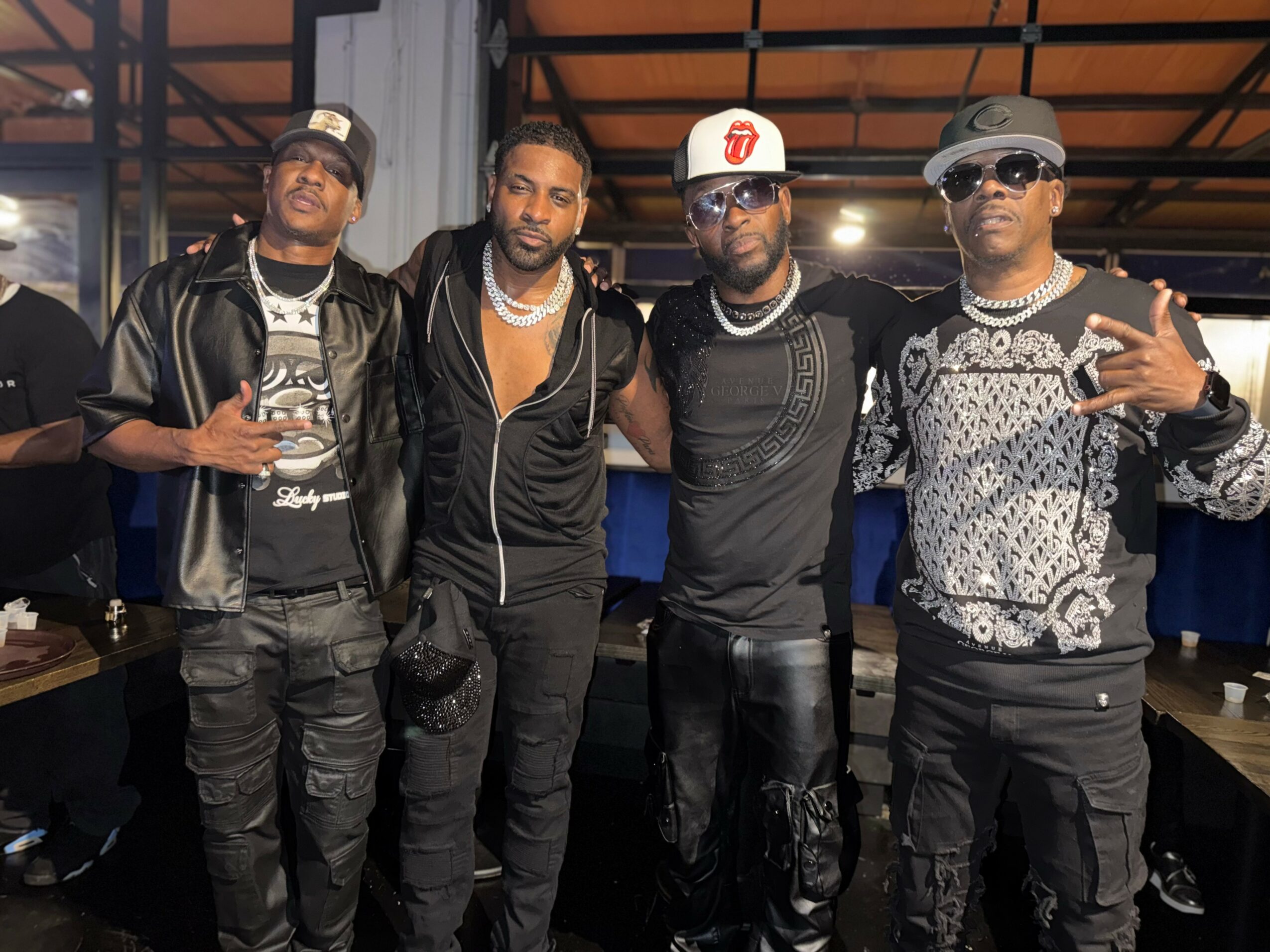 Four men standing together indoors, some wearing sunglasses and casual clothing, posing for a photo.