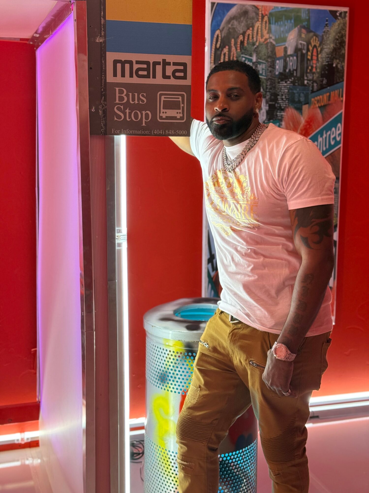 Man with beard and tattoos wearing a white t-shirt and yellow pants standing near a bus stop sign inside a colorful indoor space.