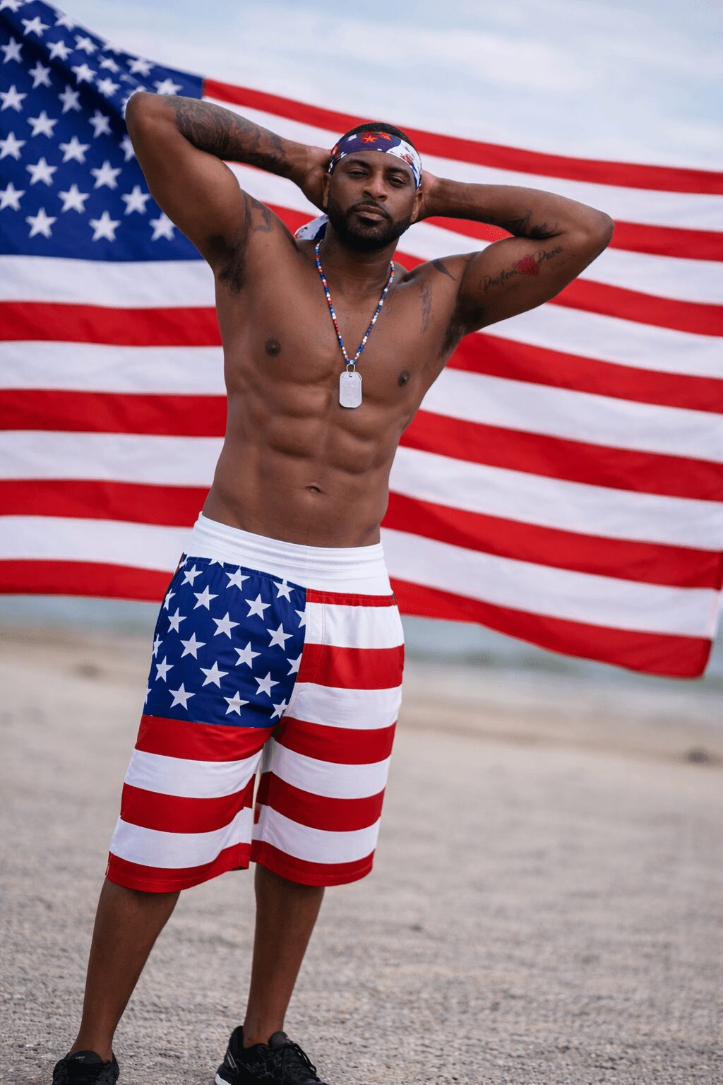 Man standing shirtless in front of an American flag, wearing patriotic shorts and sunglasses, with hands behind head.