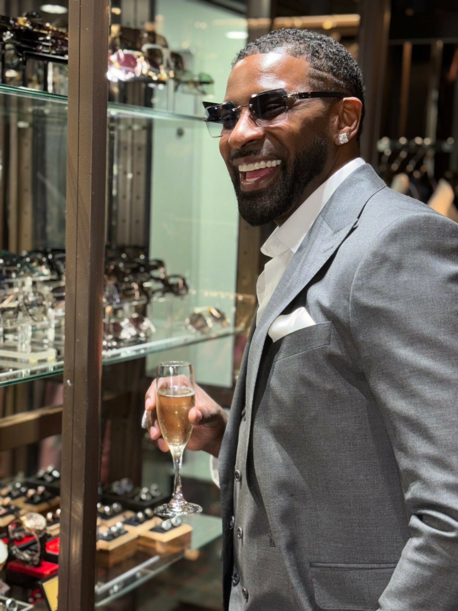 Man in gray suit and glasses holding a glass of champagne, smiling, in a store with jewelry displays.
