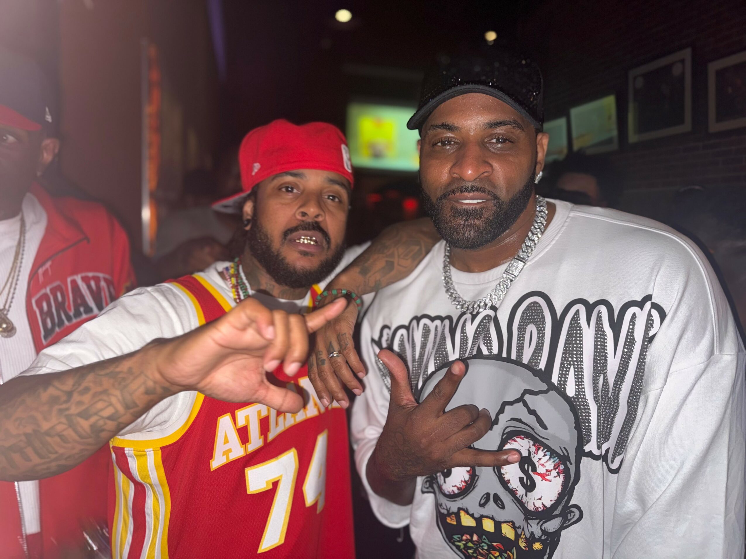 Two men pose together, one wearing a red cap and a basketball jersey, the other in a white graphic shirt, in a dimly lit setting.
