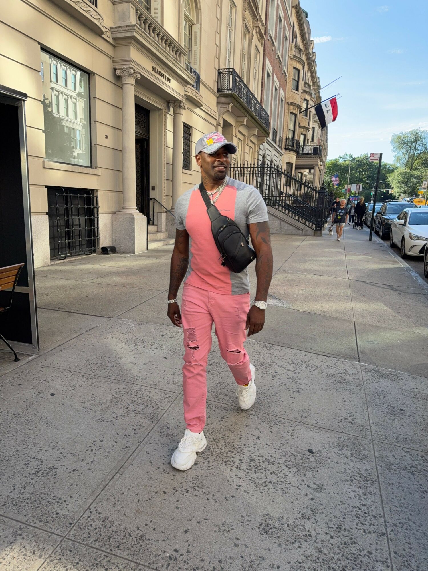 Person walking on sidewalk with buildings in background, wearing pink pants, white shoes, and a gray top, carrying a black bag.