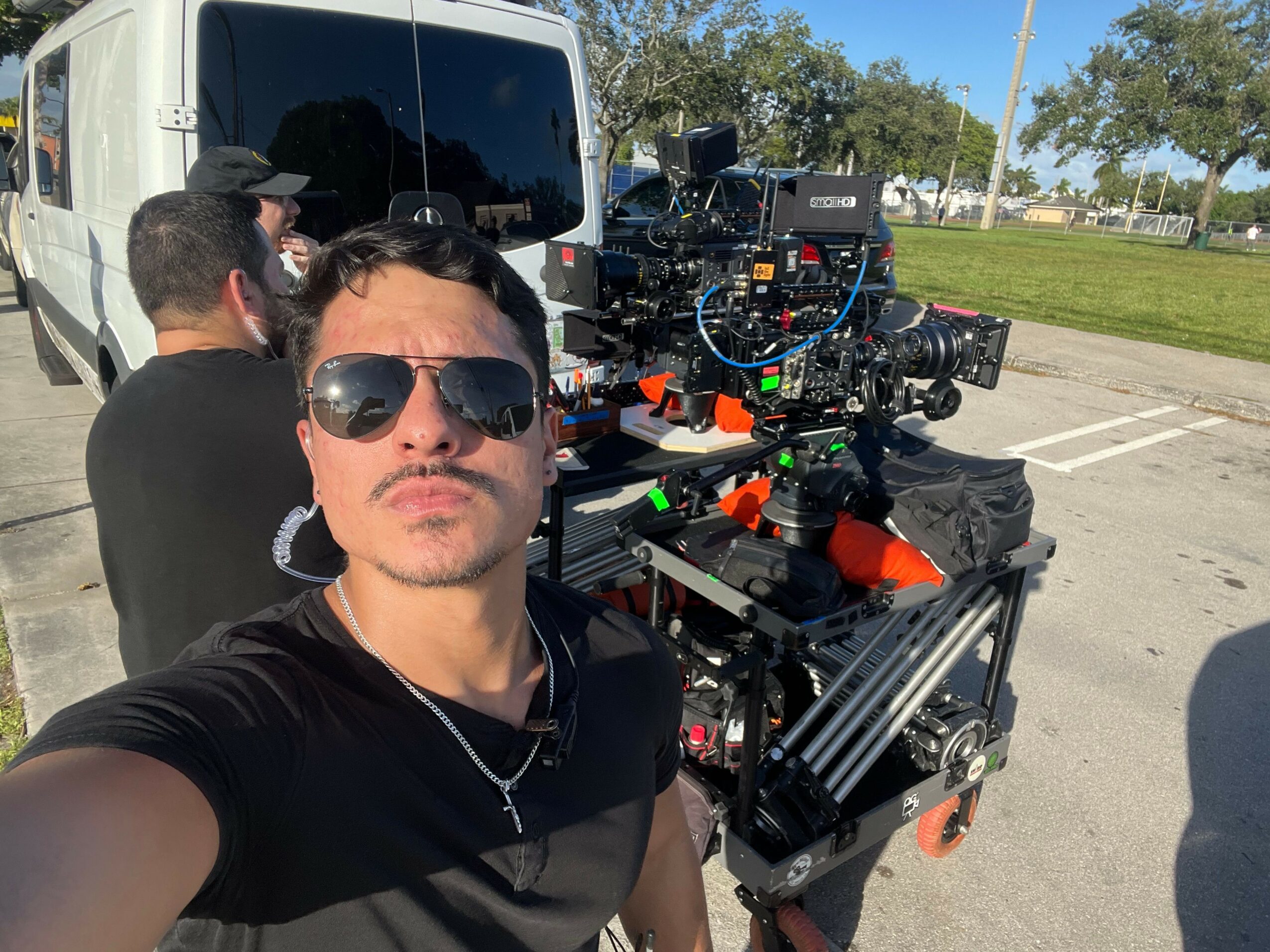 Person wearing sunglasses and black shirt taking selfie with camera equipment and vehicle in background.