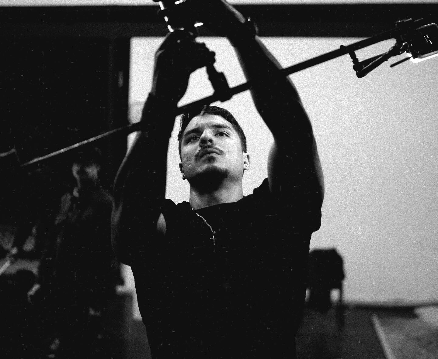 Person adjusting equipment with arms raised, looking upward, in a dark room, black and white photo.