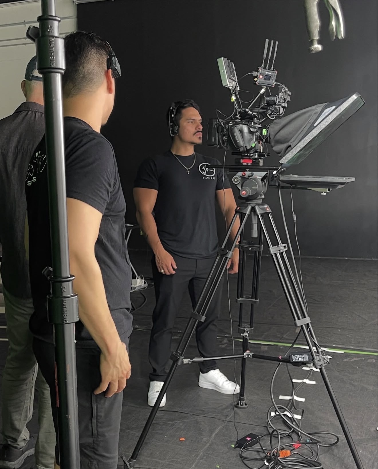 Two men on a film set with a camera on a tripod, one wearing headphones, in a dark studio.
